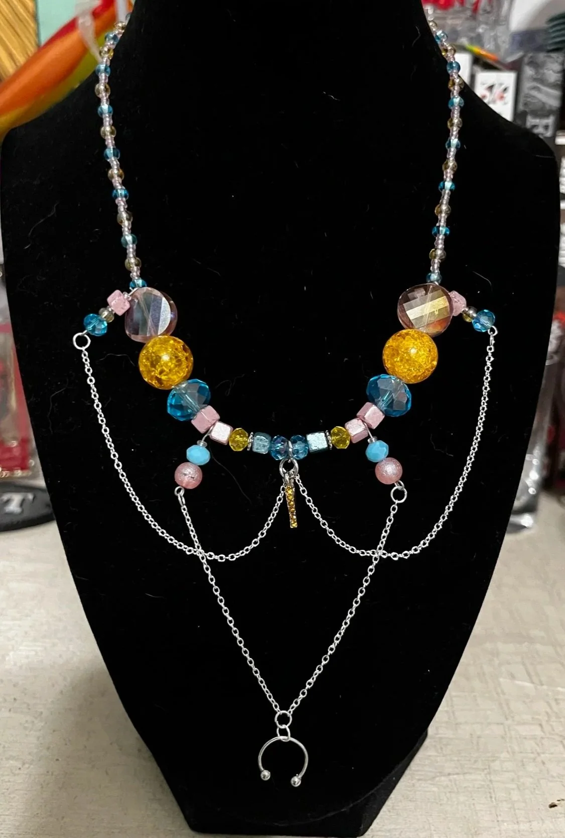 Colorful beaded necklace with yellow, blue, pink, and clear beads, silver chains, and a circular pendant on a black display stand.