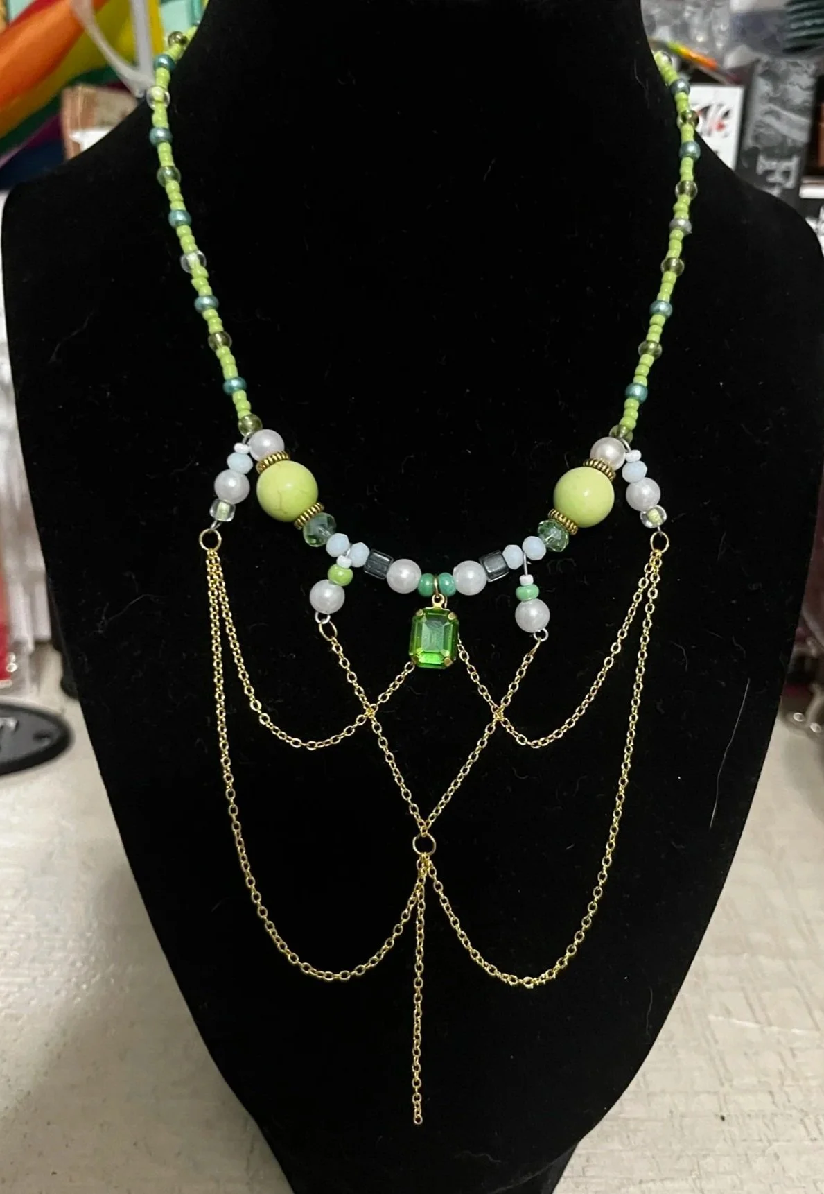 Layered necklace with green, white, and gray beads, gold chains, and a green gemstone pendant displayed on a black jewelry stand.