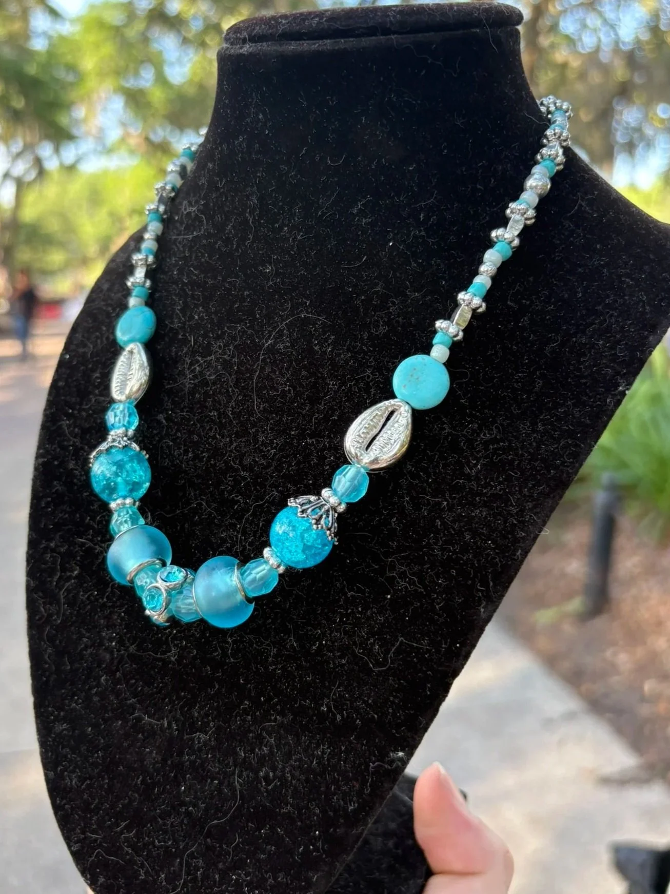 A turquoise and silver beaded necklace displayed on a black velvet necklace stand outdoors.