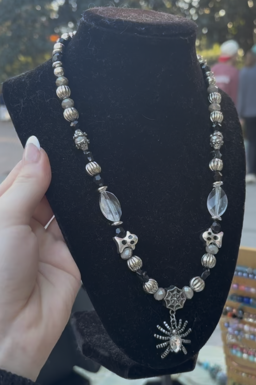 A black velvet jewelry display bust with a Halloween-themed beaded necklace featuring a spider, skulls, a web, and various beads, held by a person's hand outdoors.