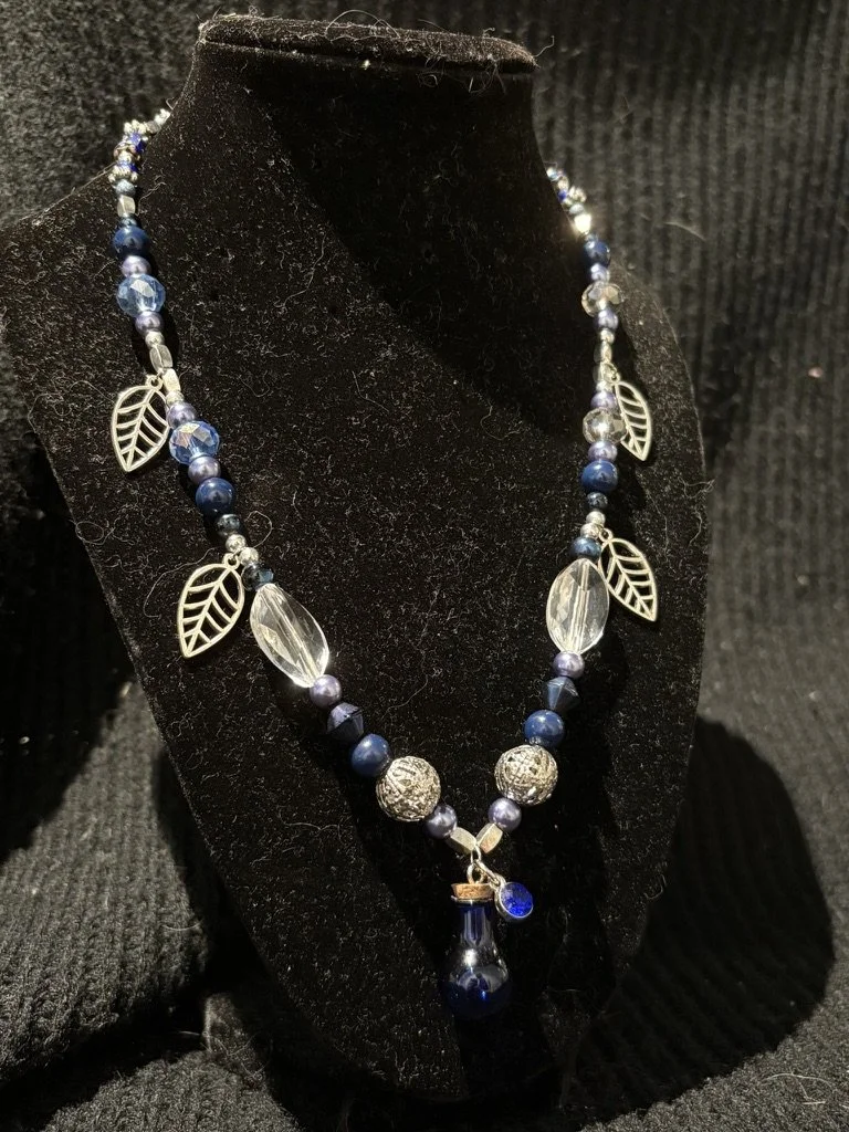 A necklace with blue and silver beads, silver leaf charms, and a blue pendant, displayed on a black velvet bust.