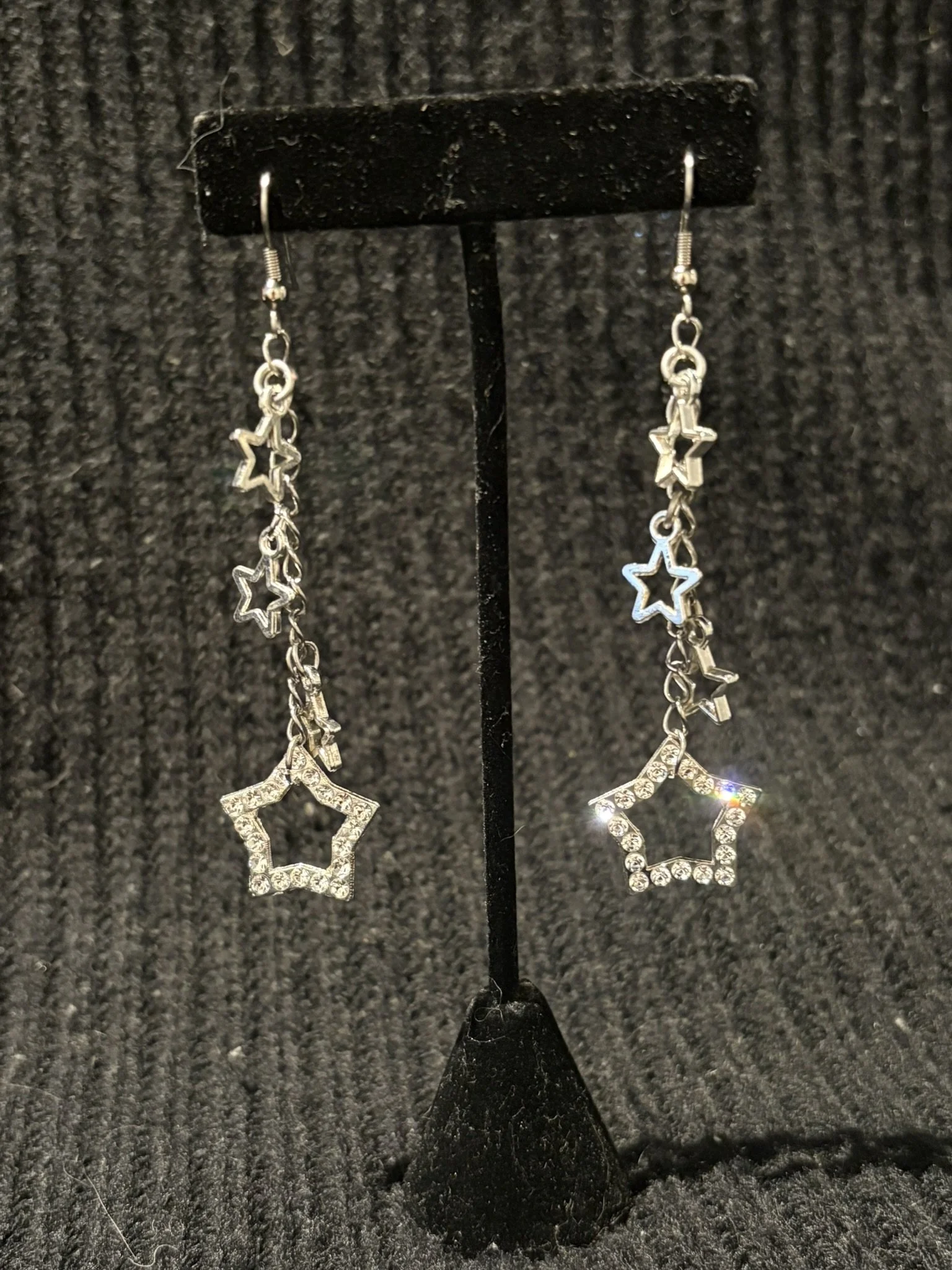 Silver star-shaped and moon-shaped dangling earrings displayed on a black stand against a dark textured background.