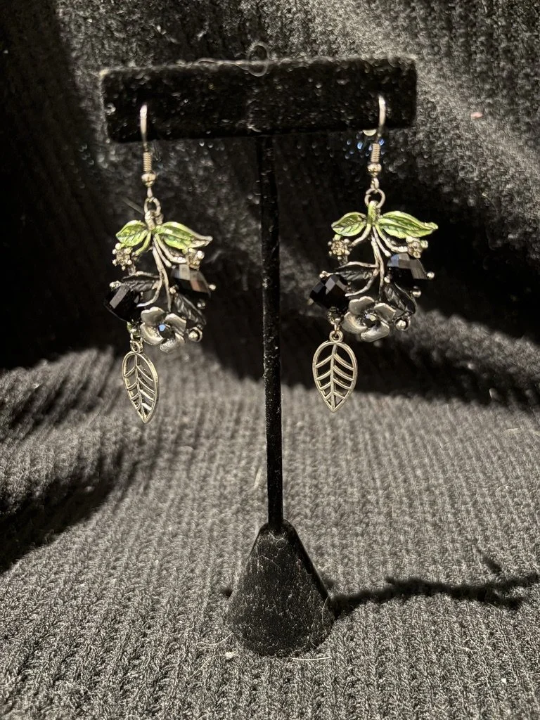 Dangle earrings with botanical leaf and berry design, featuring black and silver beads, displayed on a black jewelry stand against a black textured background.