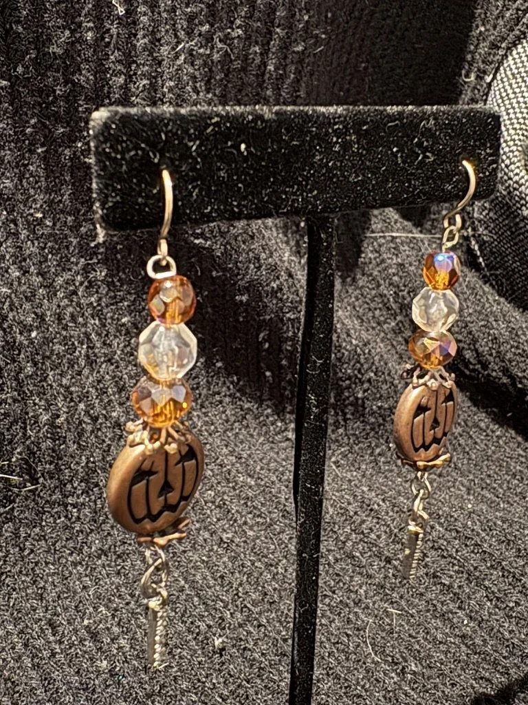 Pair of beaded earrings with amber and clear crystals, and a circular copper-toned charm with the text 'LOVE', hanging from a black jewelry display stand.