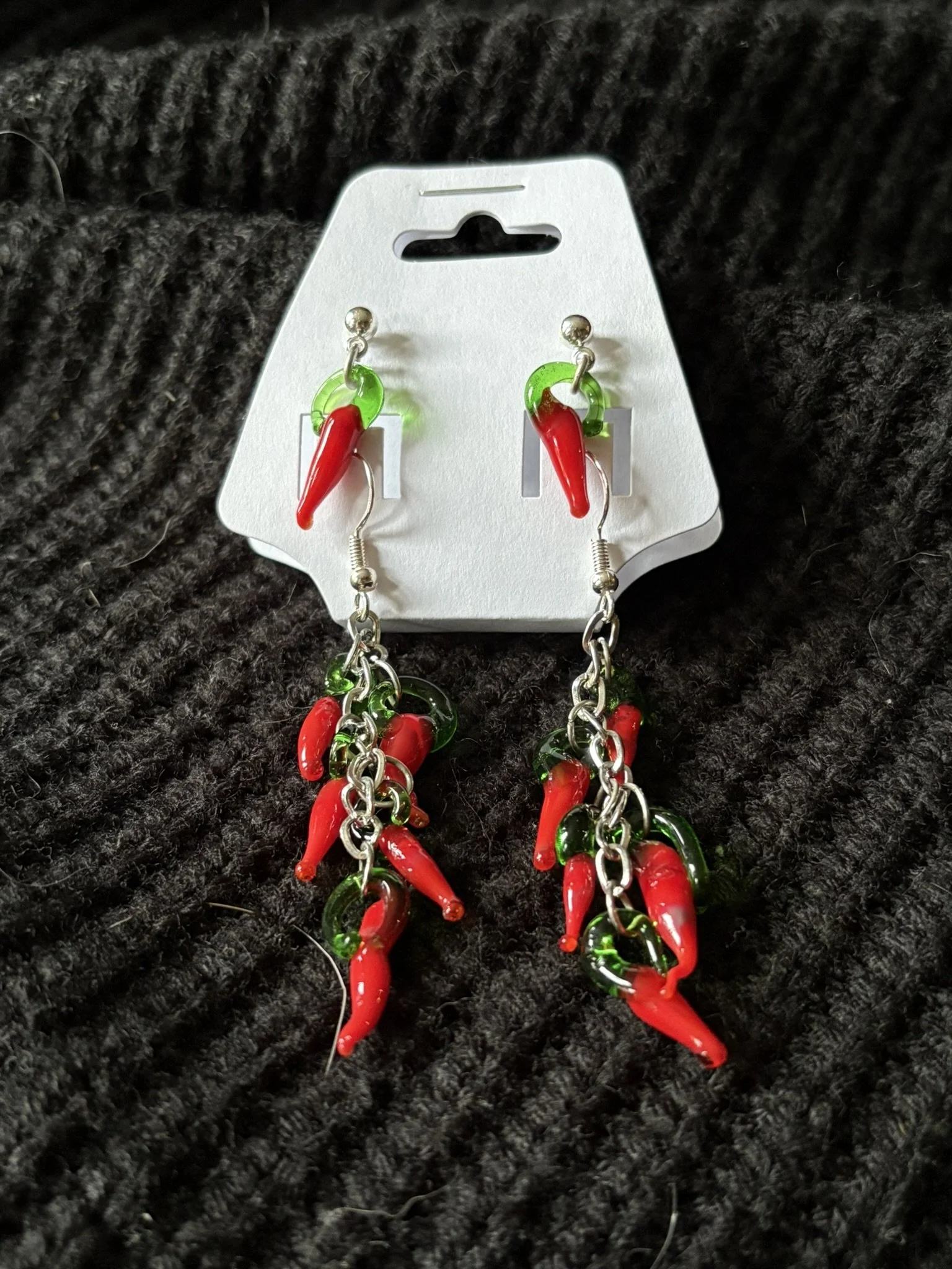 Red and green chili pepper-shaped earrings with metal chains, hanging on a white display card against a black textured background.
