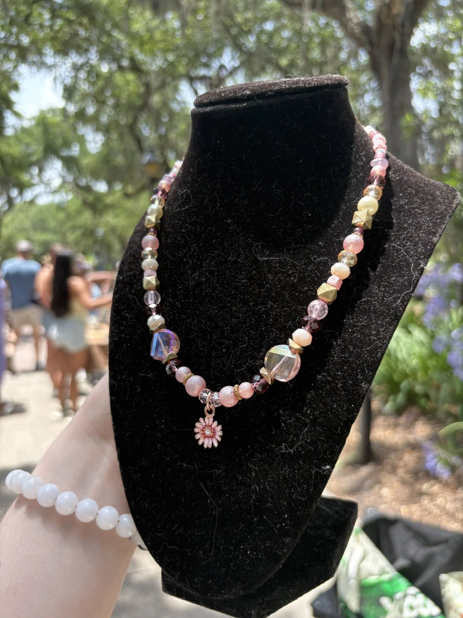 Necklace with pink, white, and gold beads and a flower pendant displayed on a black velvet bust outdoors with blurred trees and people in the background.