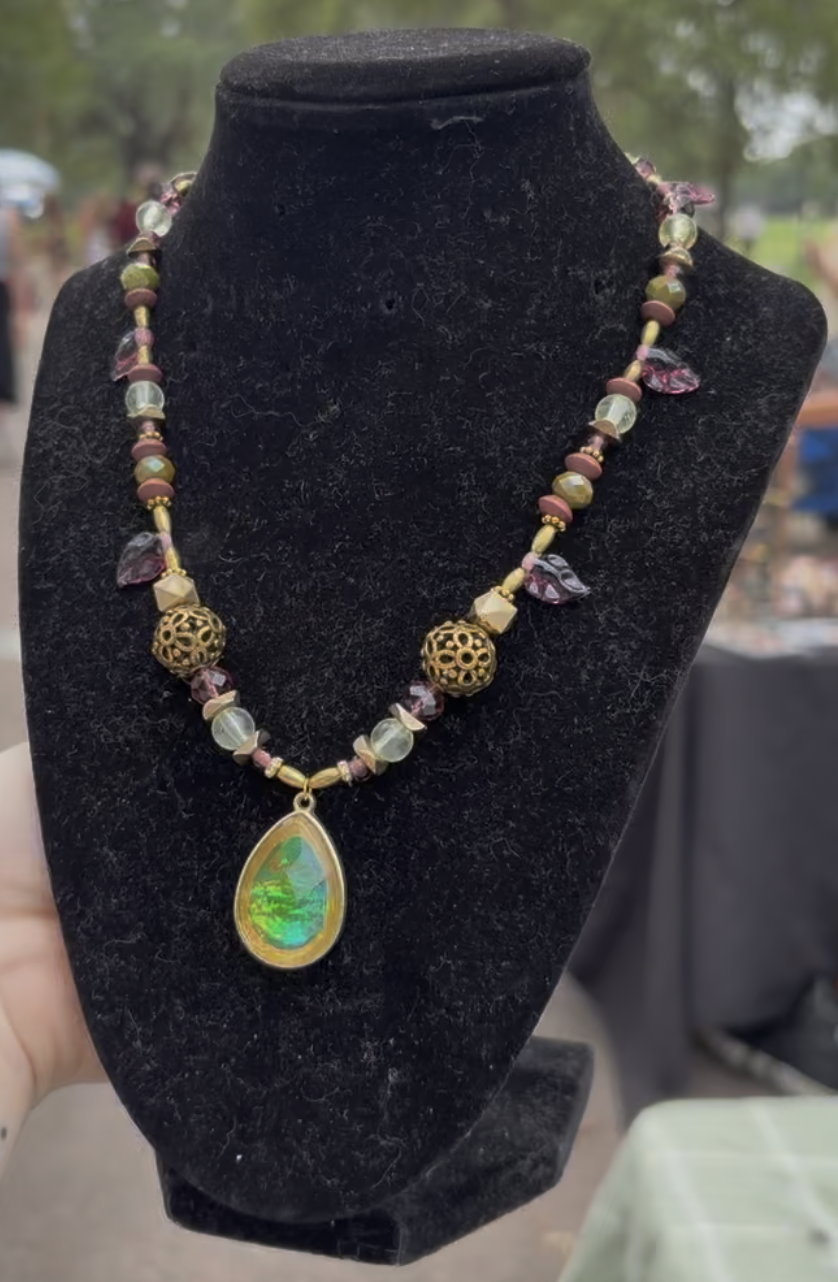Jewelry necklace with various beads, gold filigree spheres, and a green and yellow teardrop pendant displayed on a black stand