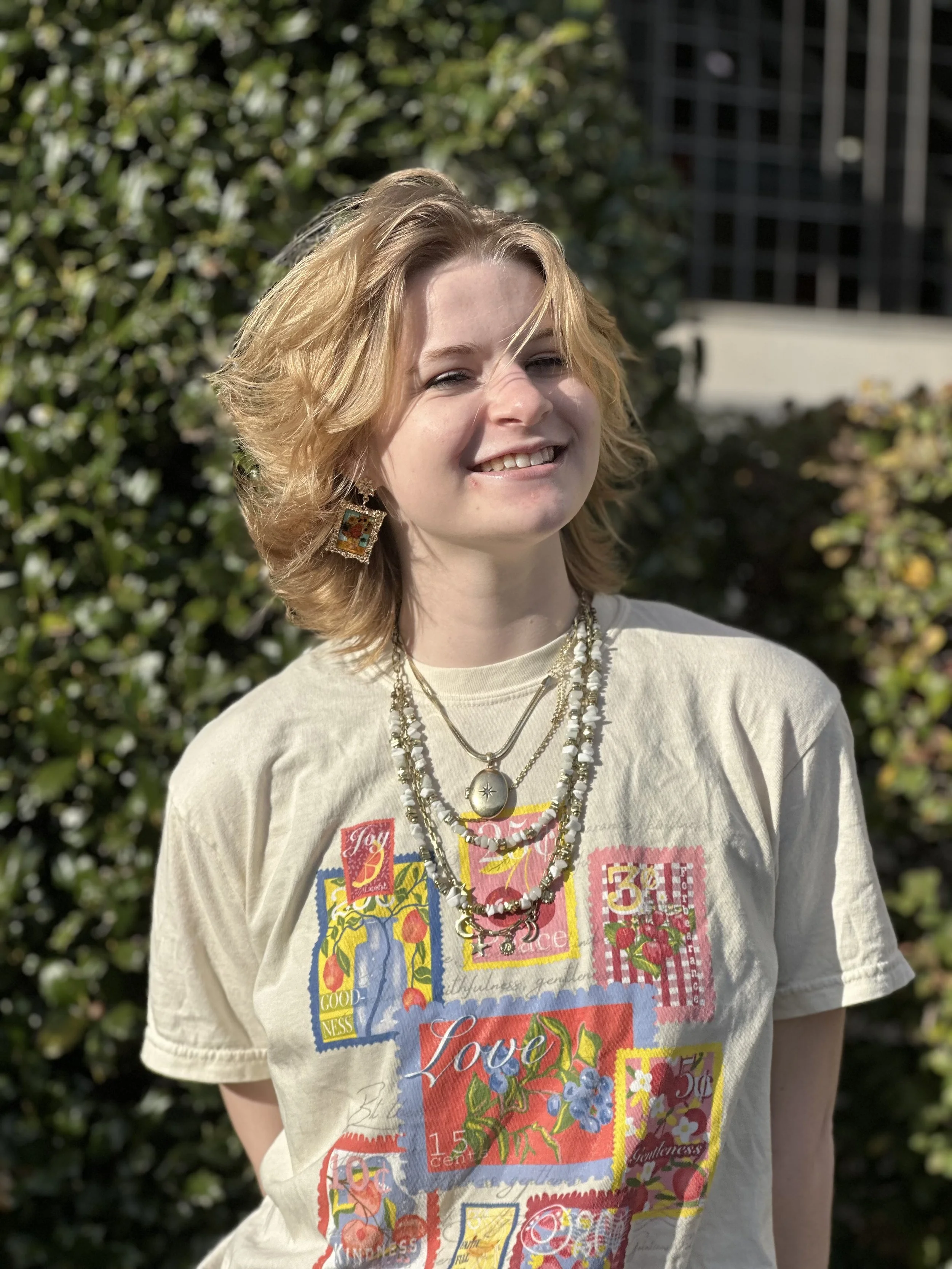 A woman with blonde, shoulder-length hair smiling outdoors, wearing colorful jewelry and a T-shirt with a vibrant, collage-style graphic design featuring words like 'Love' and 'Goodness', against a background of trees and buildings.
