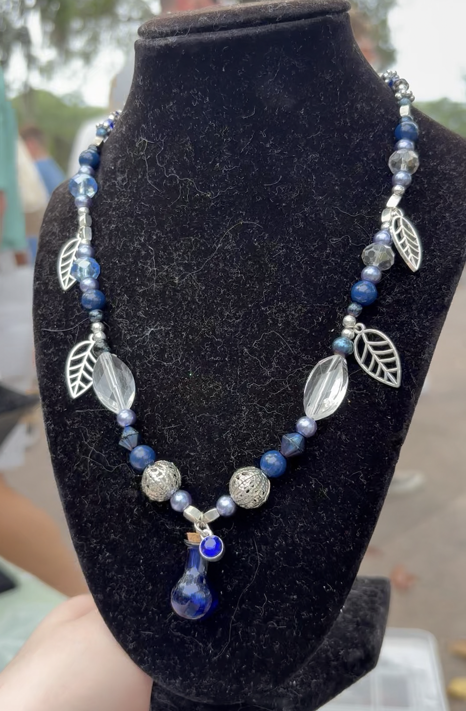 Blue and silver beaded necklace with leaf and crystal charms displayed on a black velvet necklace stand.