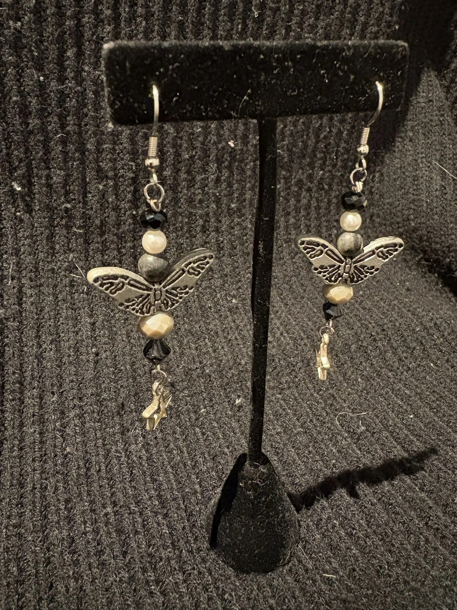 A pair of dangling earrings with butterfly and moon designs, beads, and metal charms, displayed on a black textured surface.