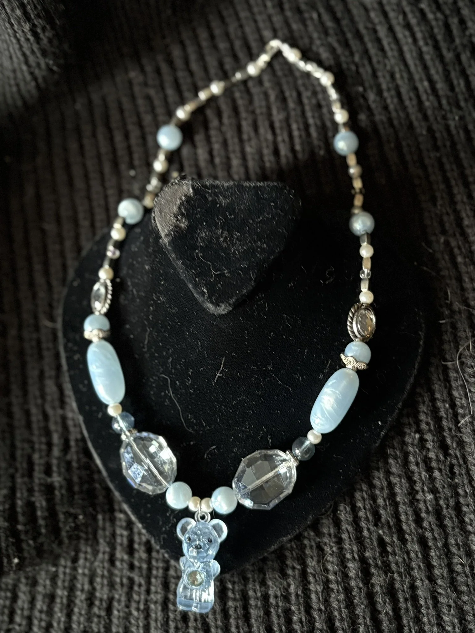 A necklace with large clear and light blue beads, a bear-shaped pendant, and a figured crystal pendant, displayed on a black velvet bust with a textured black background.