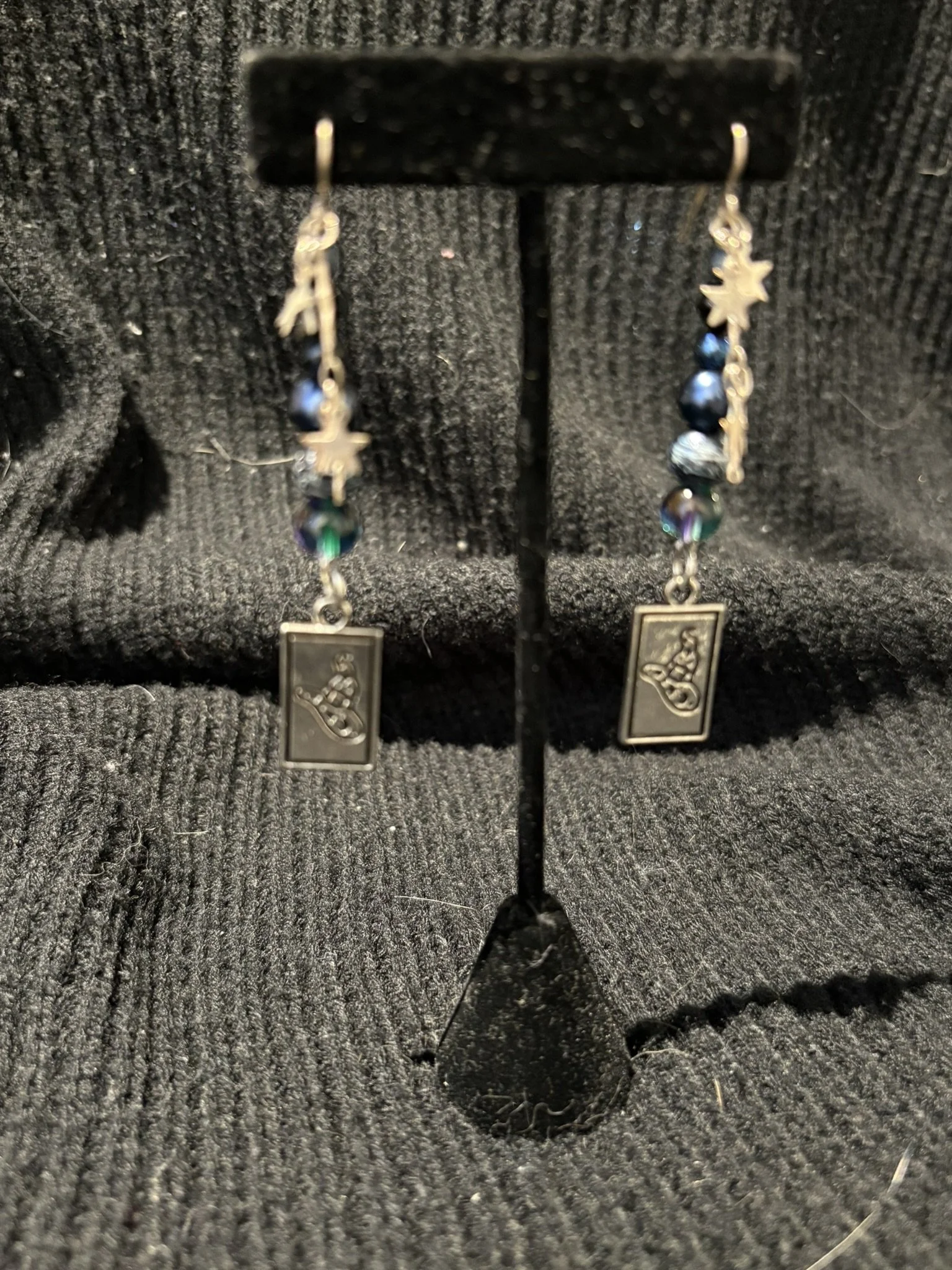 A pair of dangle earrings with black beads and metal charms resting inside a black jewelry box on a textured surface.