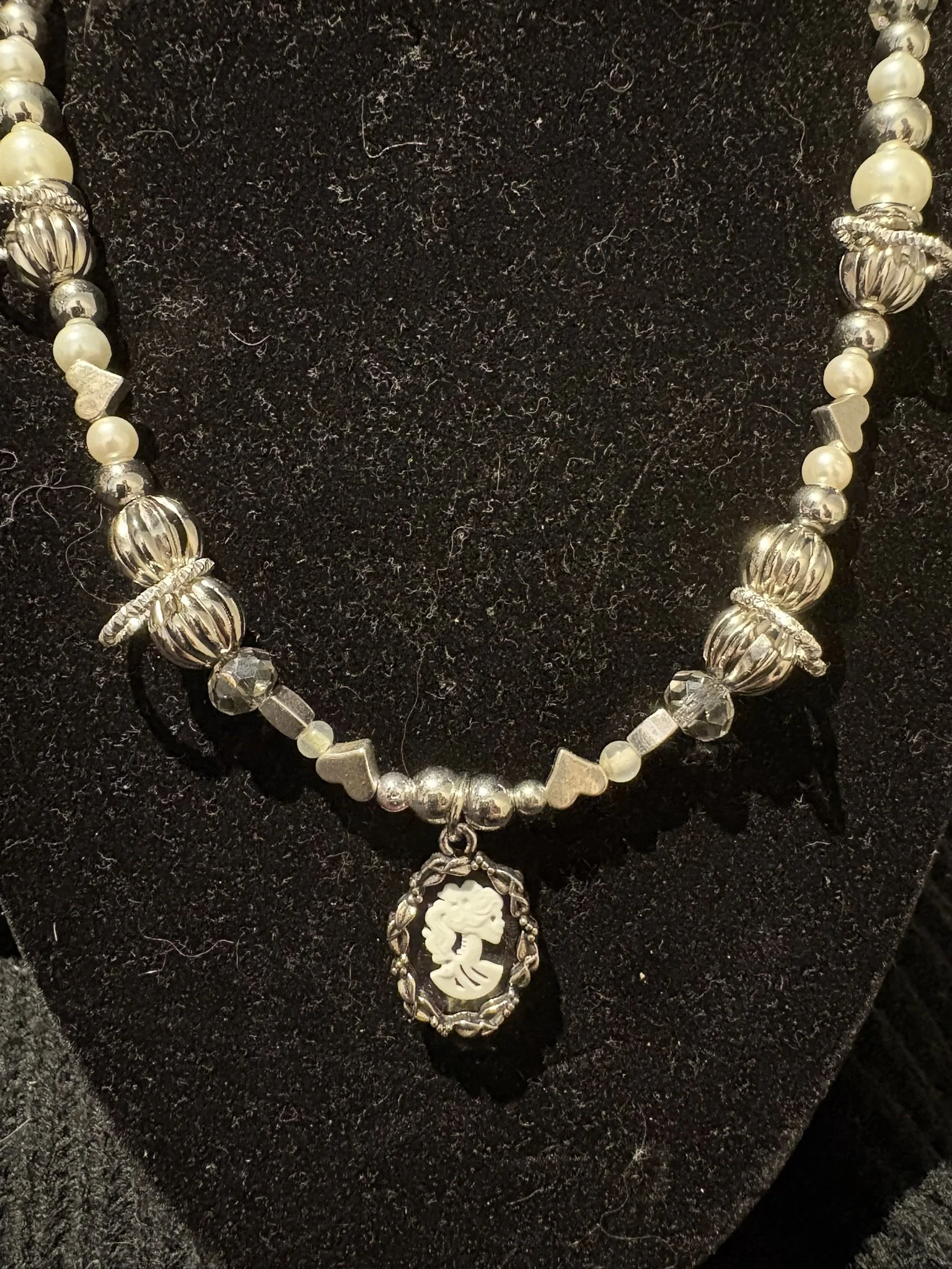 A pearl necklace with large silver beads, a flower-shaped pendant, and a carved white pendant hanging from it, placed on a black textured fabric