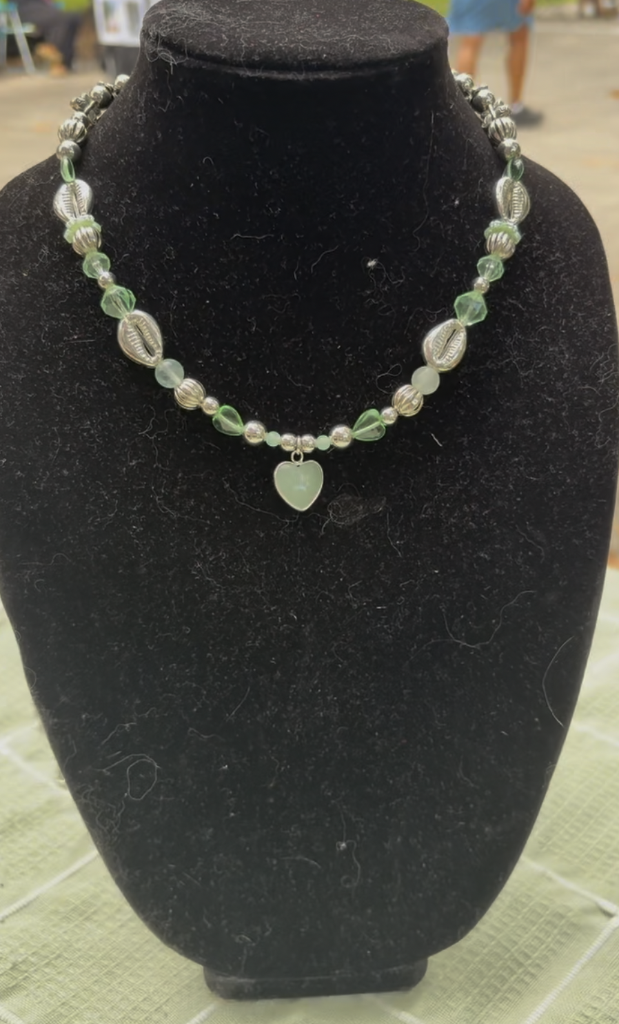 Beaded necklace with green, silver, and silver cowrie shell beads and a heart-shaped pendant, displayed on a black velvet bust.