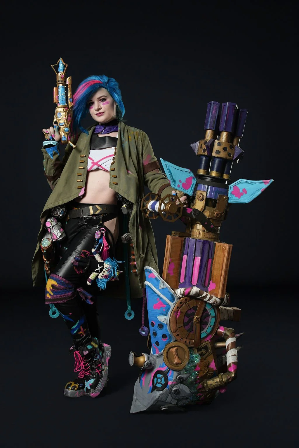 A woman dressed as a colorful, futuristic warrior with bright blue hair, holding a large, decorated weapon and wearing a green jacket, black shorts, and vibrant accessories, standing against a black background.