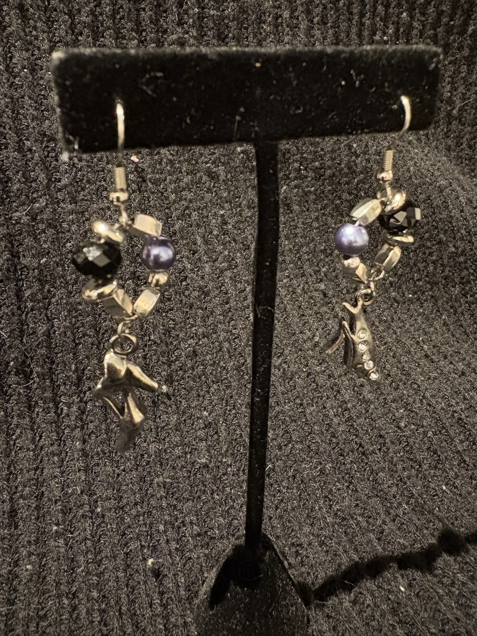 A pair of dangling earrings with dark purple pearls and metallic beads, hanging from a black display stand on a dark, textured surface.