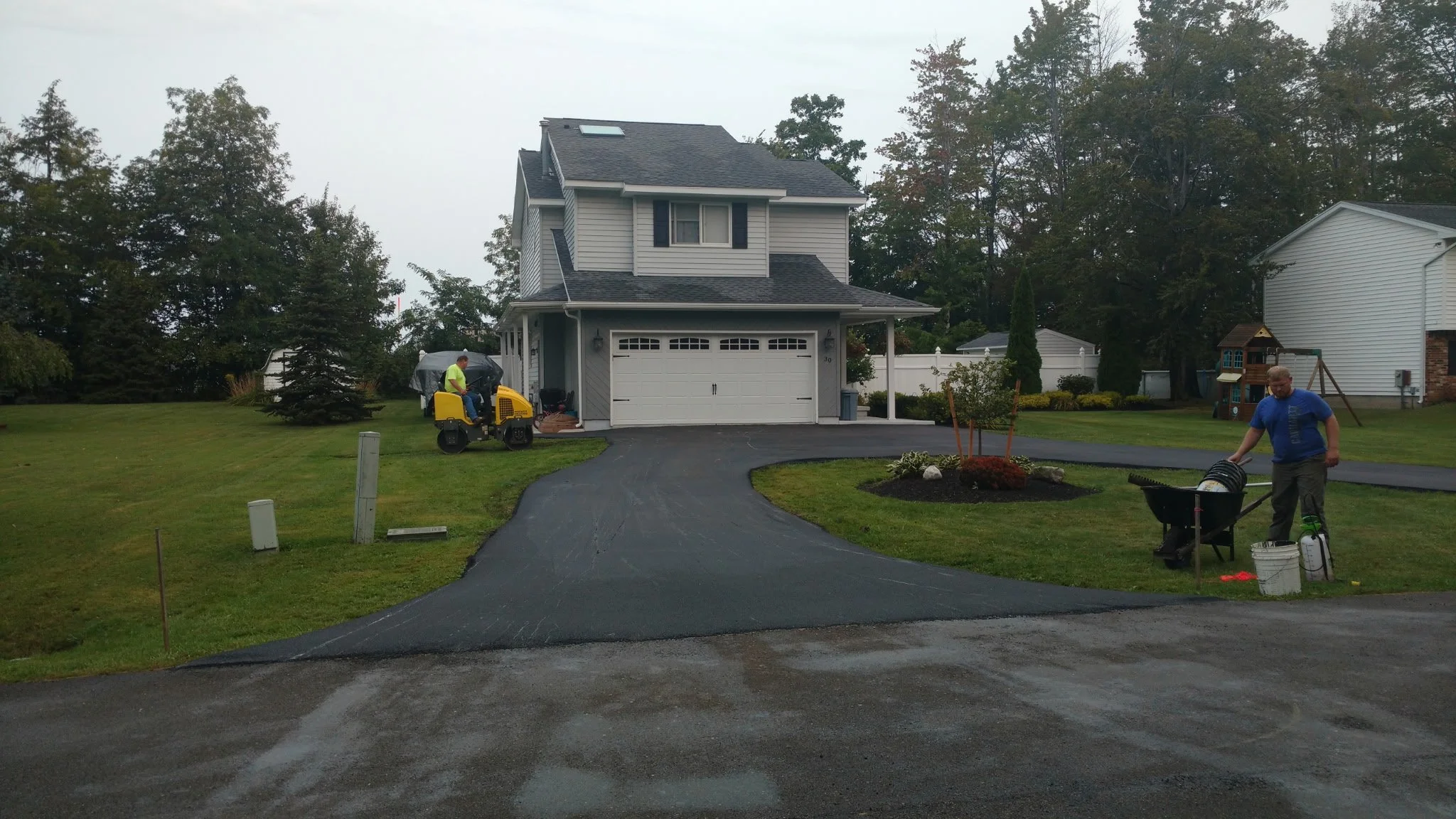 Two workers are paving a driveway in front of a two-story house during overcast weather. One worker is operating a compactor, and the other is managing equipment near a wheelbarrow.