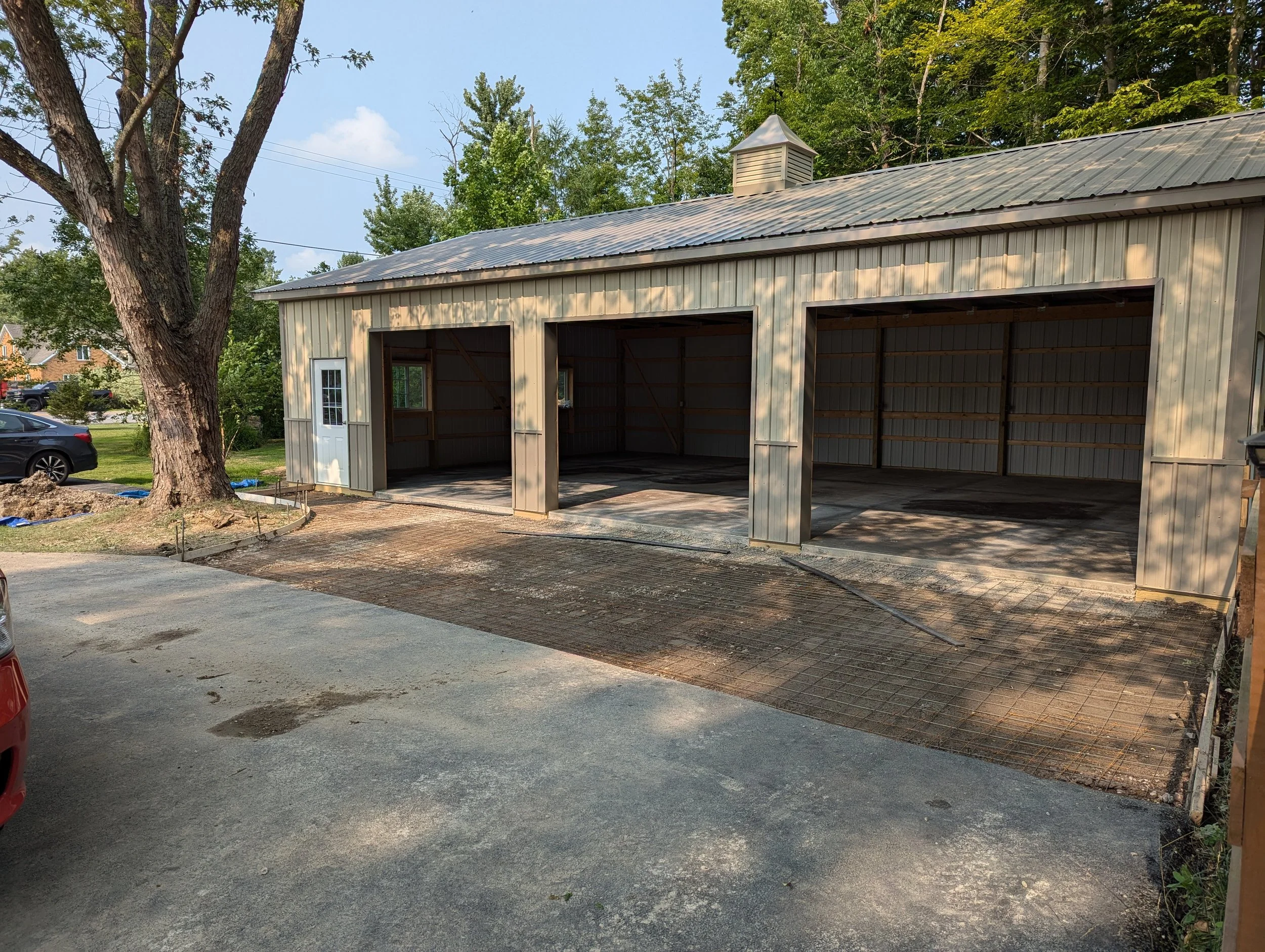 A partially built metal garage with three bays, set in a residential area. The garage has a metal roof and siding, with a small white door on the left side. The driveway is under construction with some gravel and dirt.