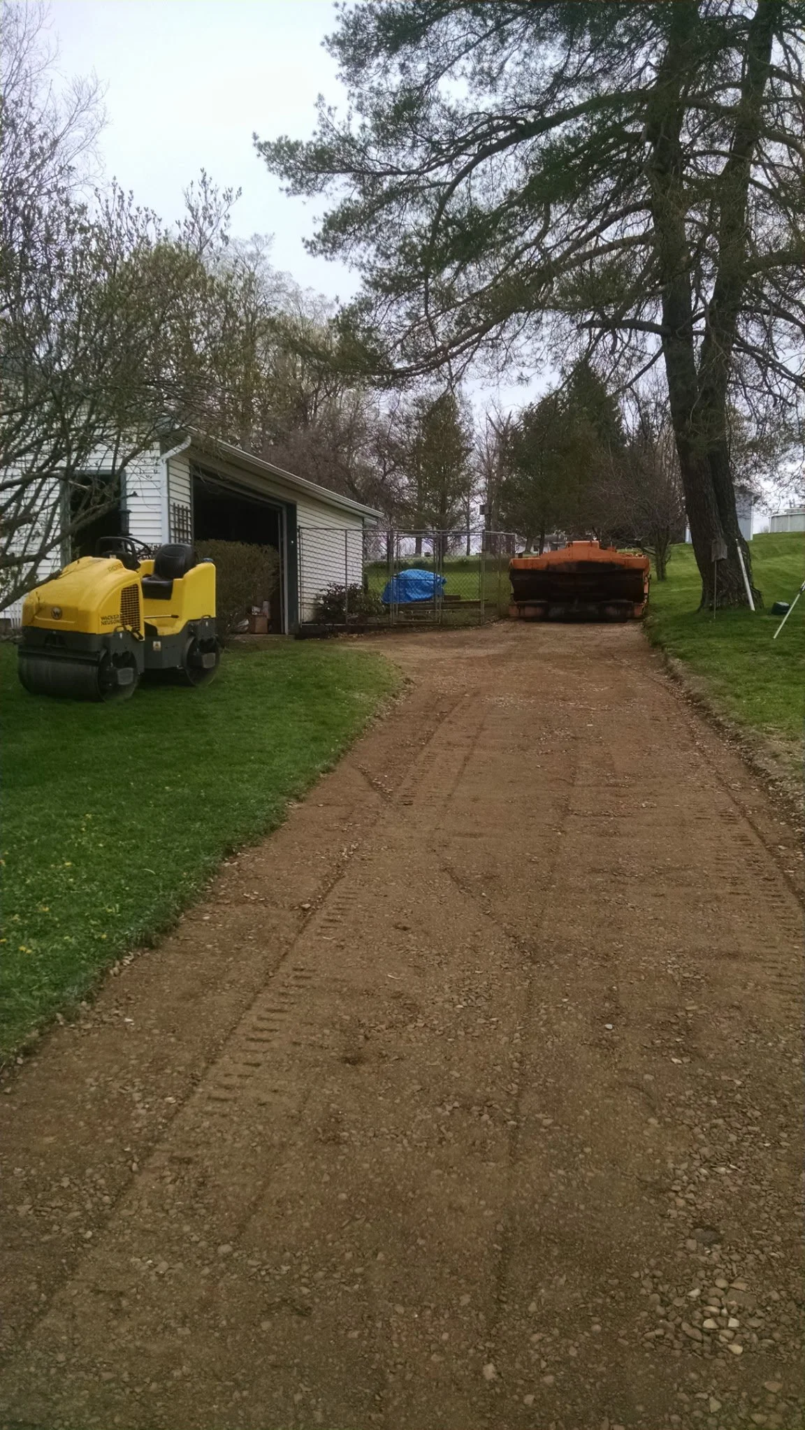 Dirt driveway with tire marks, next to a small patch of grass, leading to a fenced work area with construction equipment and trees in the background.