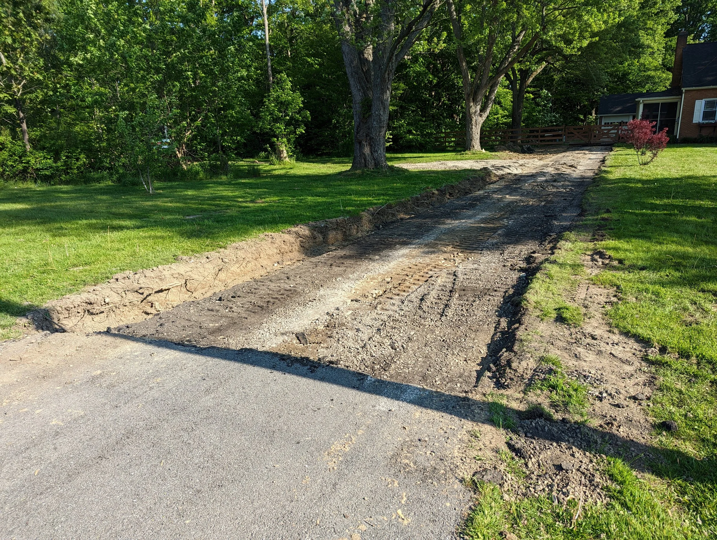 A driveway under construction, partially excavated with dirt and gravel, leading up to a house with a green lawn and trees in the background.