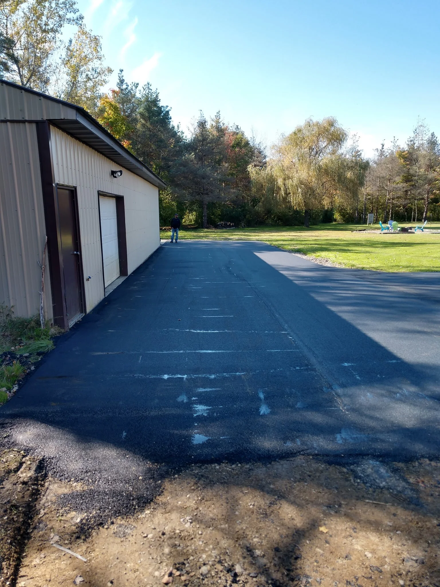 Freshly paved black asphalt driveway next to a light-colored metal building with a white garage door, surrounded by a grassy yard with trees in the background and a person standing near the building.