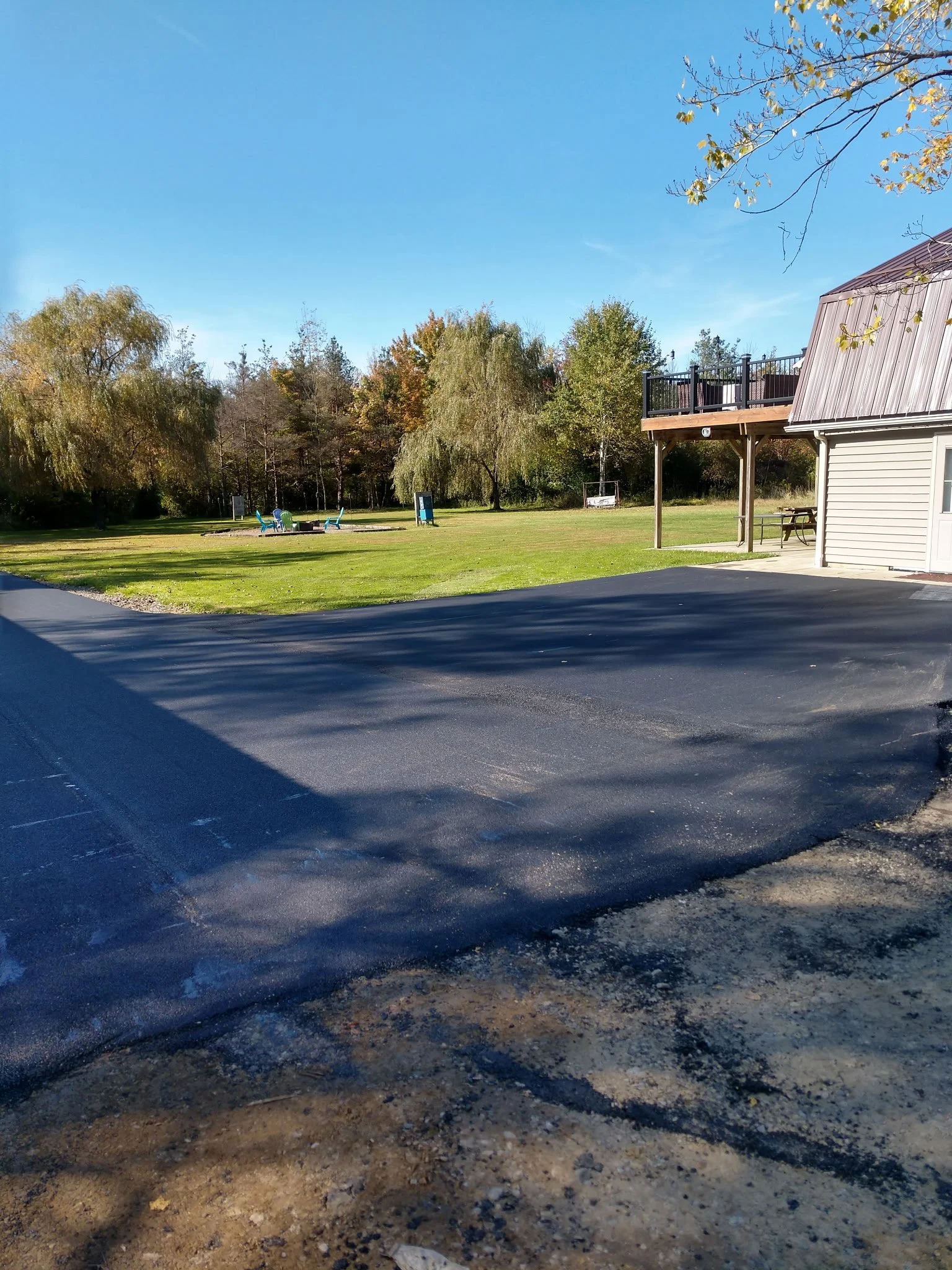 A newly paved black asphalt parking lot in front of a grassy field with trees and some outdoor seating. A building with a deck is on the right side, and the sky is clear and blue.