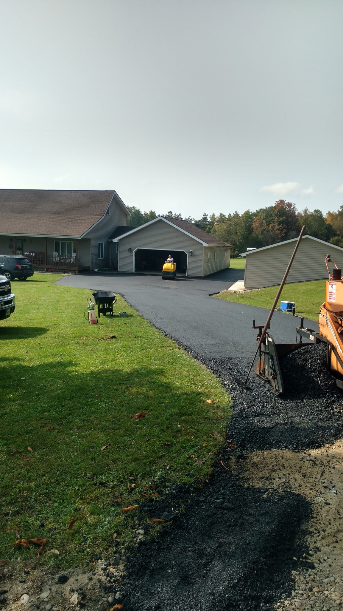 A driveway under construction with fresh asphalt, a small black wheelbarrow, and construction equipment, with a house and garage in the background.
