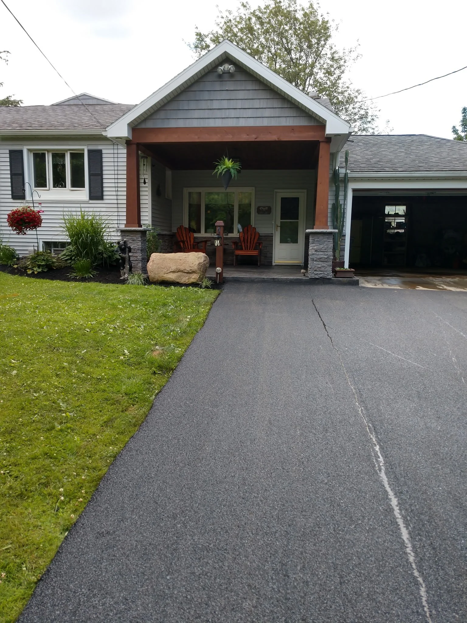 Front view of a house with a dark asphalt driveway leading to a covered porch area with outdoor chairs and a hanging plant, surrounded by greenery and a lawn.