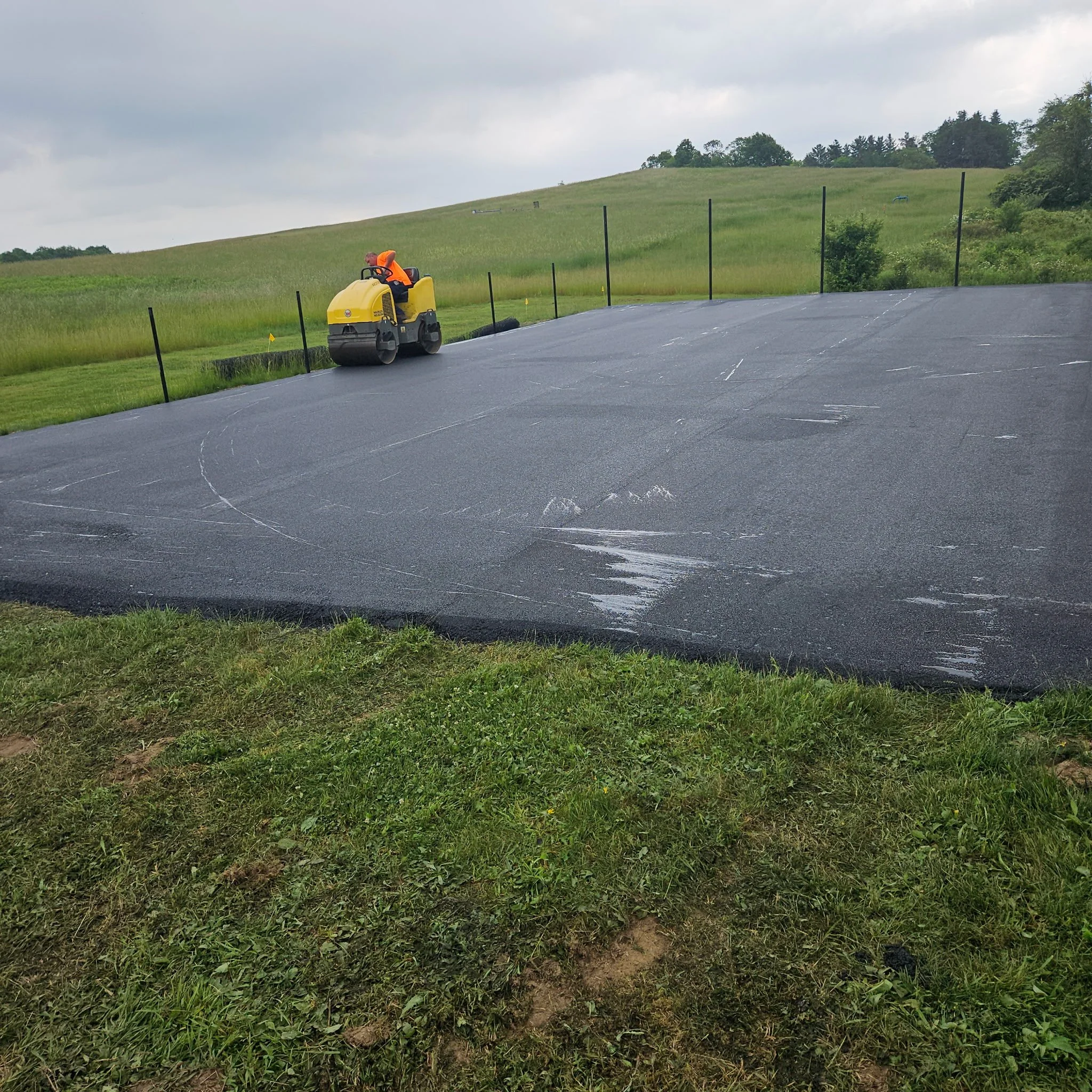 A worker operating a roller compactor on a new asphalt surface in a grassy field, with a hill and cloudy sky in the background.
