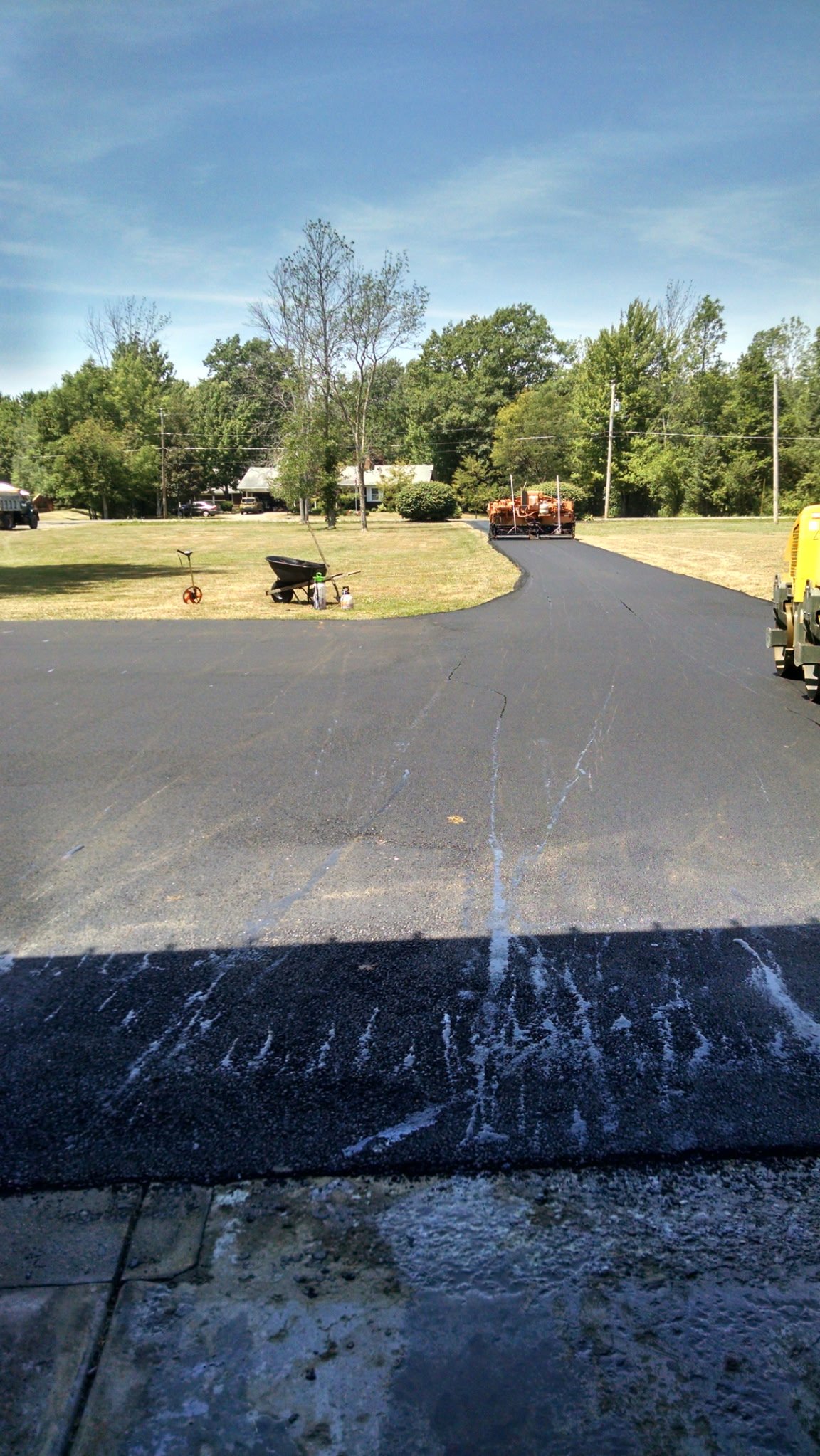 View of a newly paved driveway with construction equipment, including a roller, and a garden wheelbarrow on a grassy yard with trees and houses in the background.