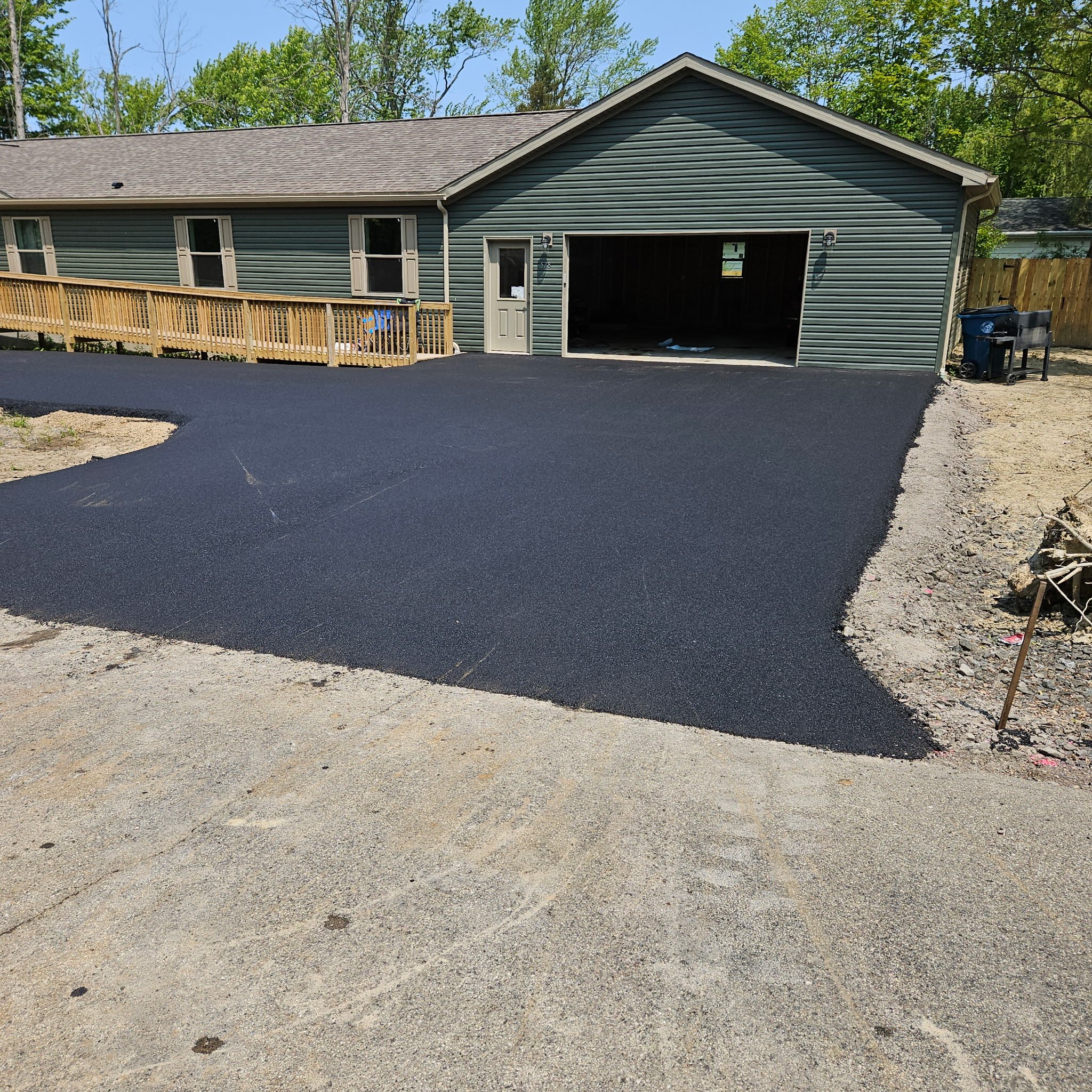 Recently paved black asphalt driveway leading to a garage with a green exterior and a white door, surrounded by partially completed gravel and dirt ground.