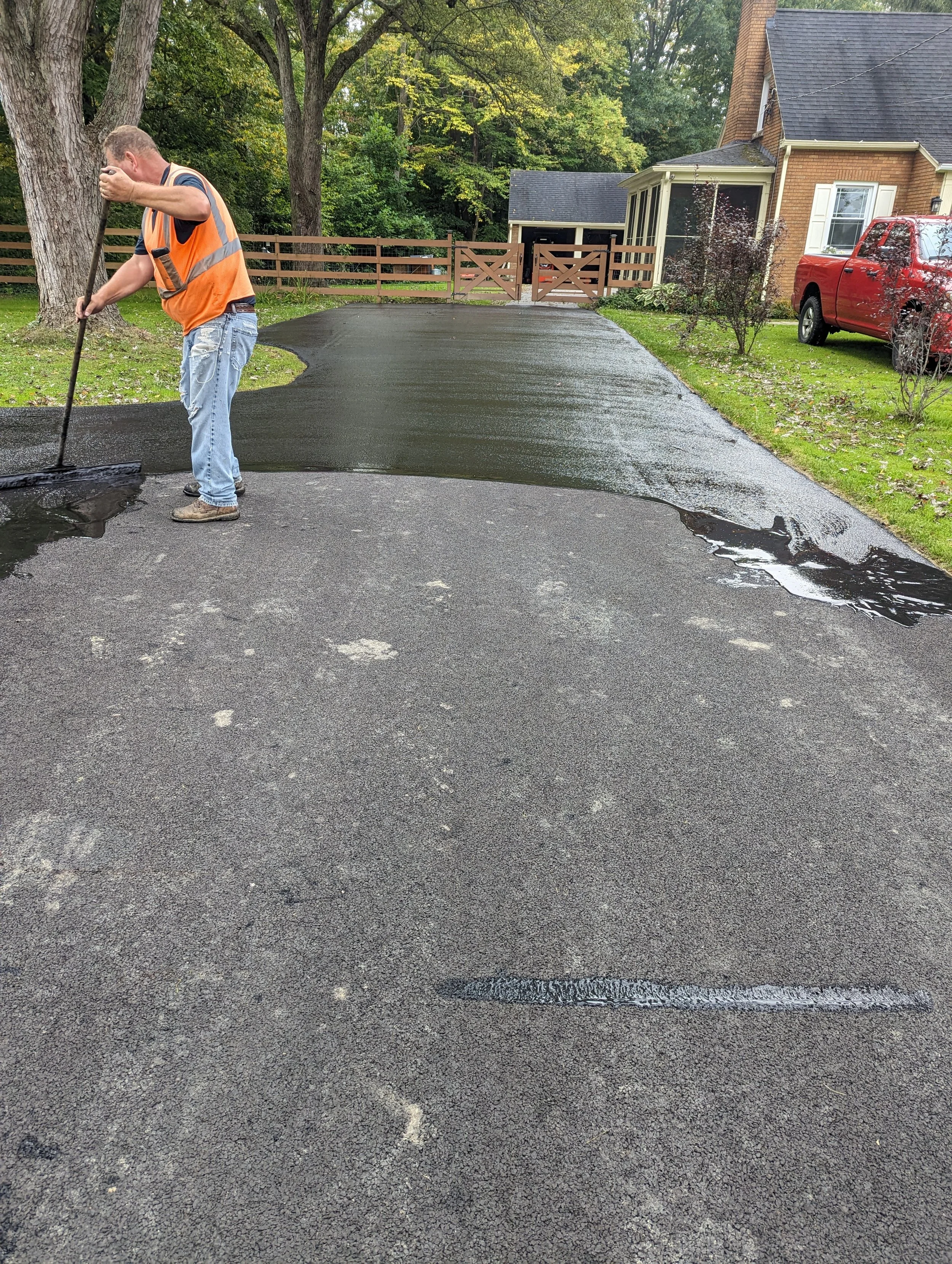 A worker in an orange safety vest and jeans is sealing a driveway with hot asphalt. The driveway leads to a house with a red pickup truck parked nearby, surrounded by trees and greenery.