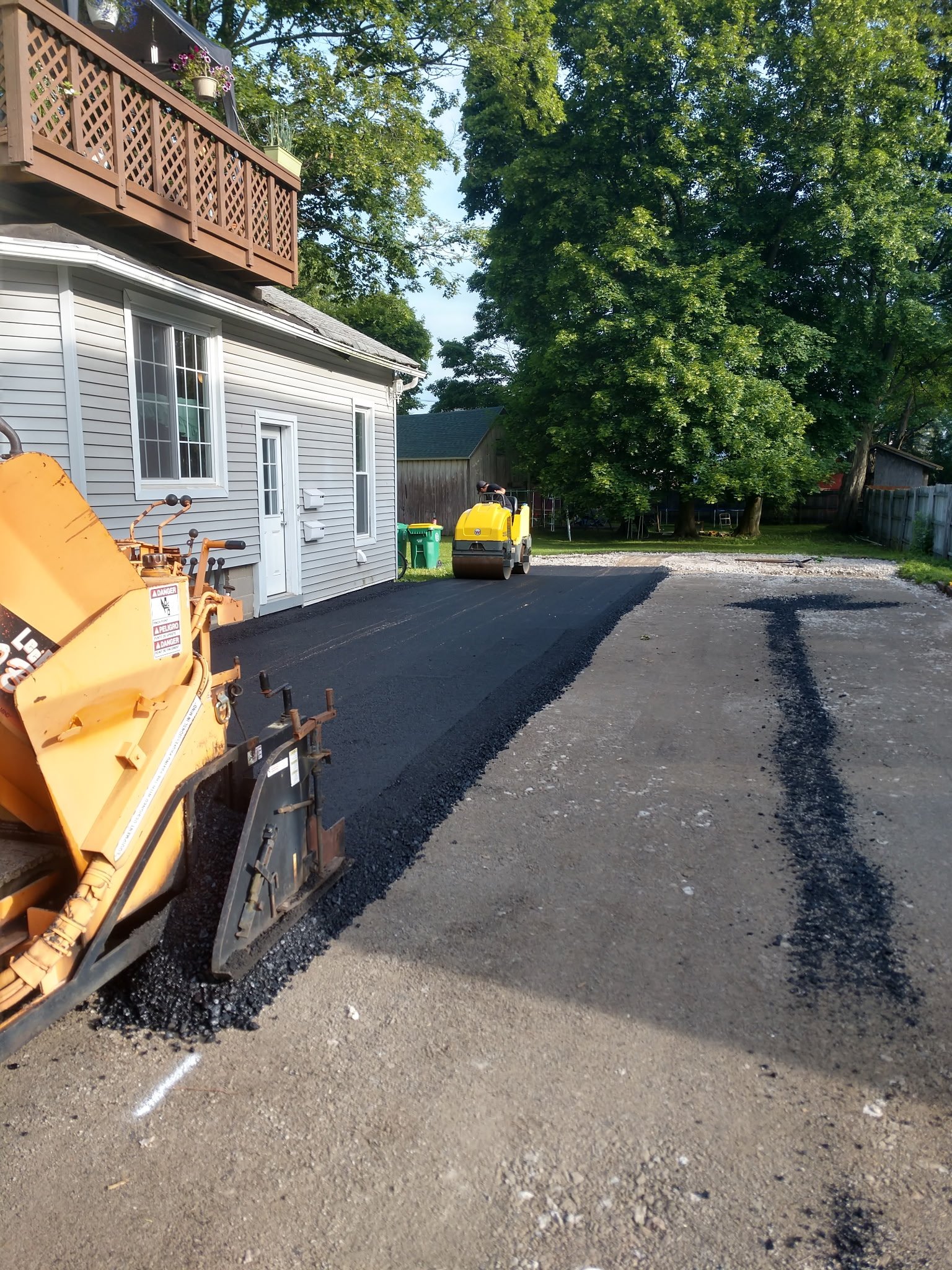 A driveway under construction with asphalt being laid down and a roller compactor in the background. There is a house with a balcony and trees in the background.