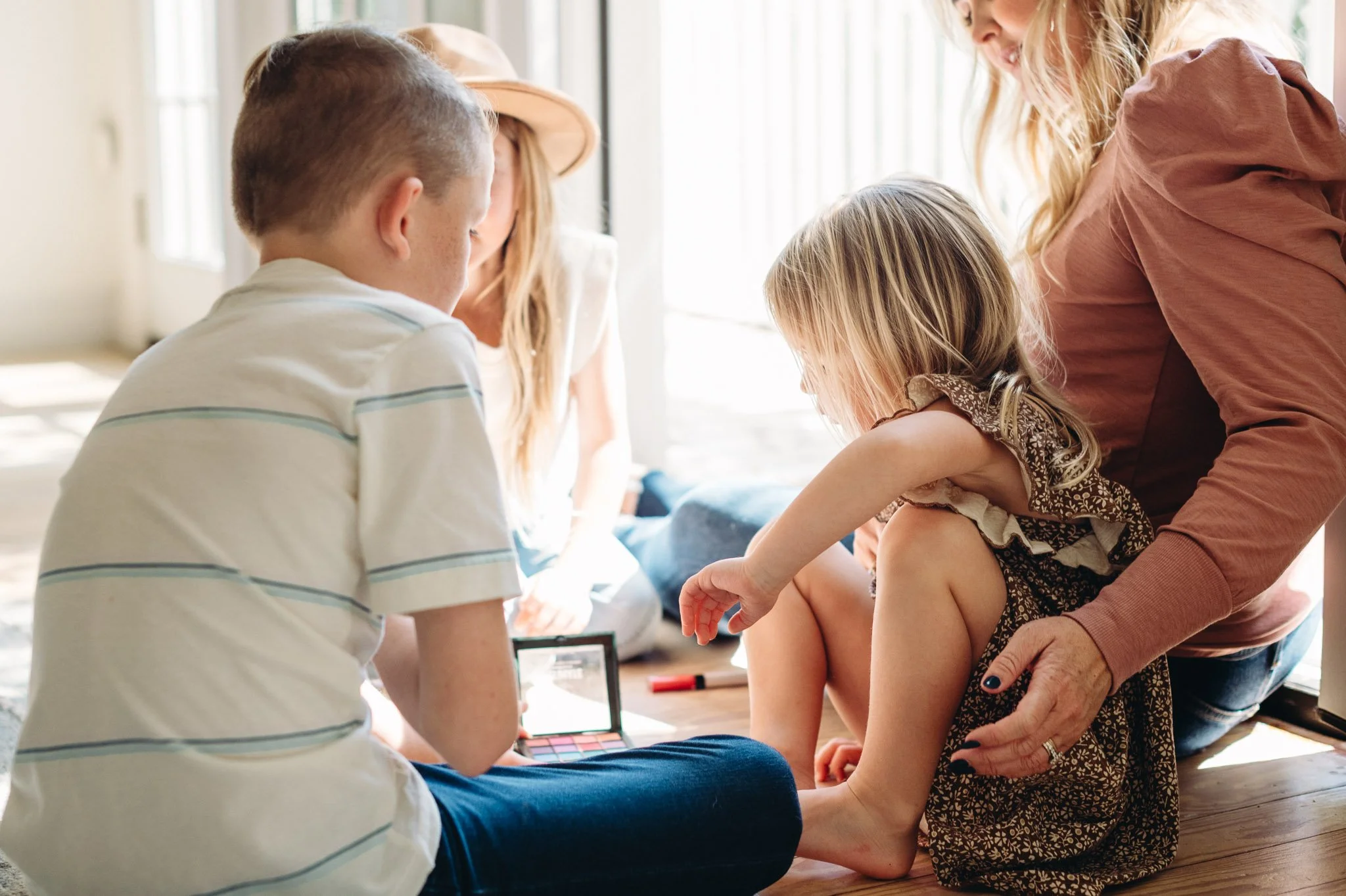 A woman with blonde hair and a pink shirt is helping three young siblings play with makeup in documentary family photo by Elise Souders.
