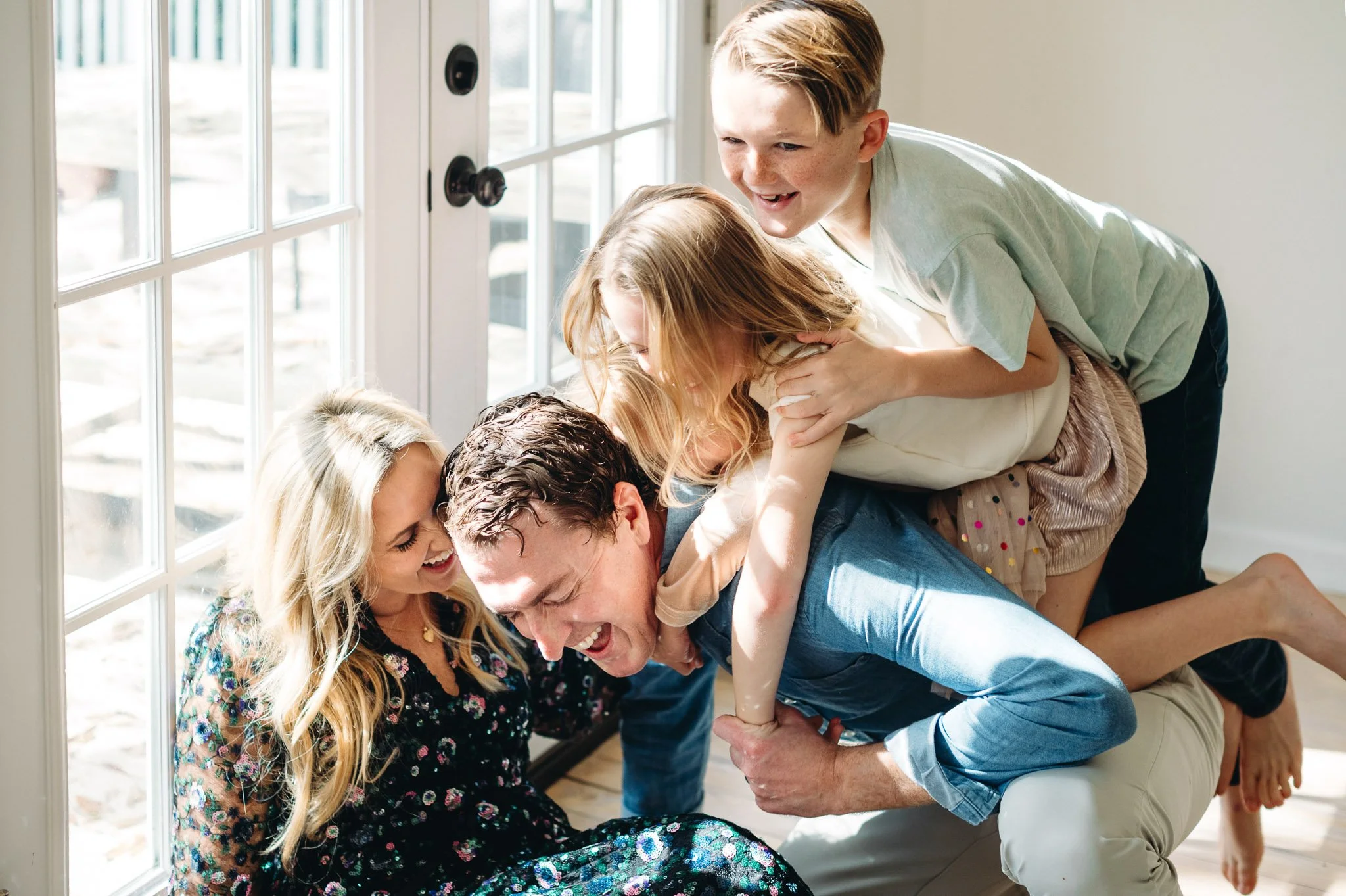 Children piled onto their father's back in a sunlit house, as father and mother laugh together by the window in-home family photography Bend, Oregon.