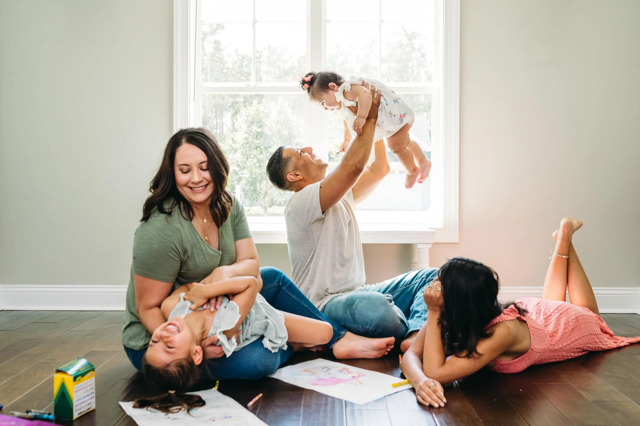 A family plays in a sunlit room of their Bend, Oregon home with crayons on the floor, dad lifting baby, and mom tickling toddler as an older sibling looks on.