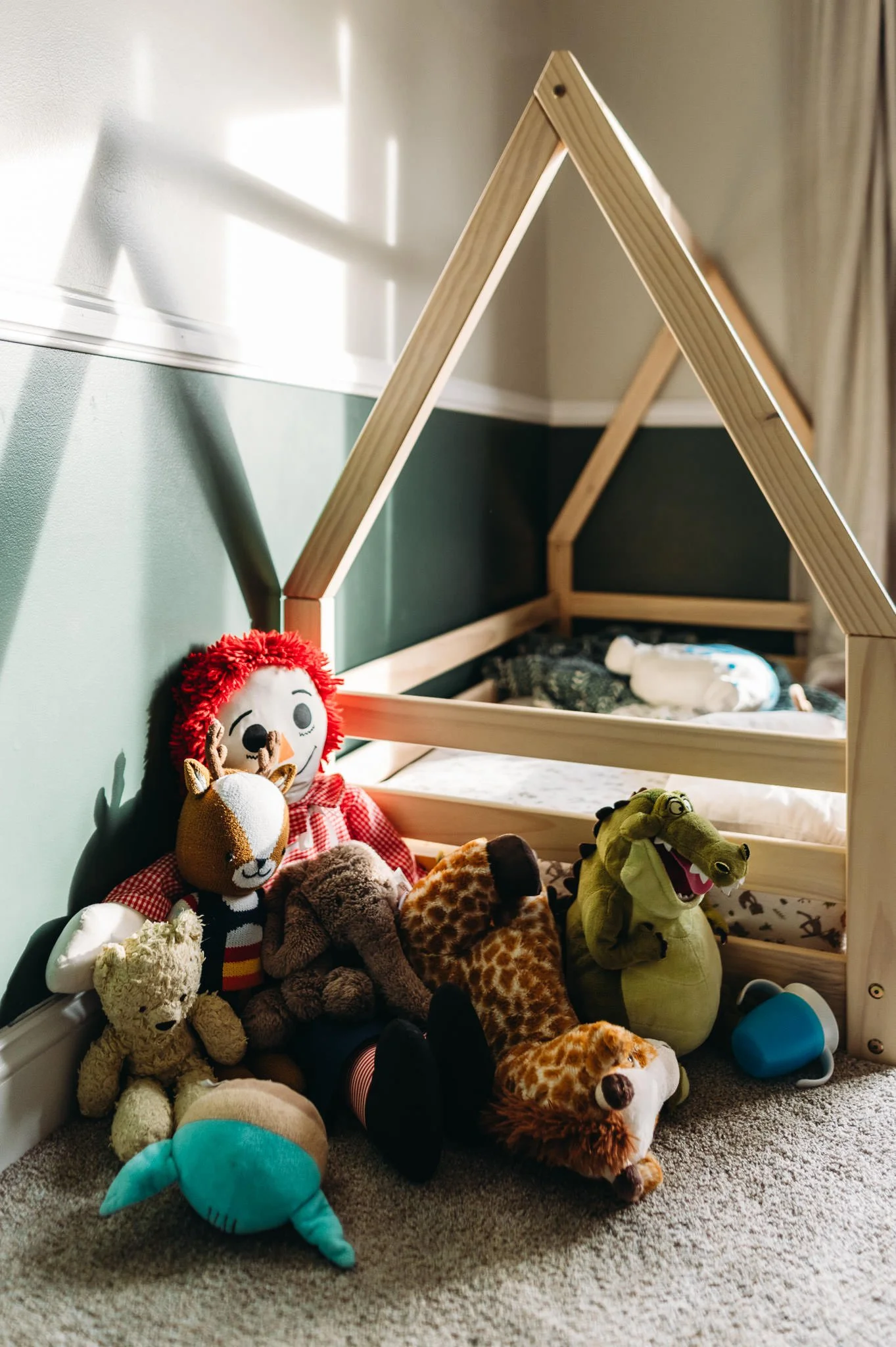 A child's bedroom corner with a wooden house-shaped bed frame, stuffed animals including a teddy bear, giraffe, crocodile, lion, hedgehog, and a plush doll, and a blue sippy cup on a beige carpet.
