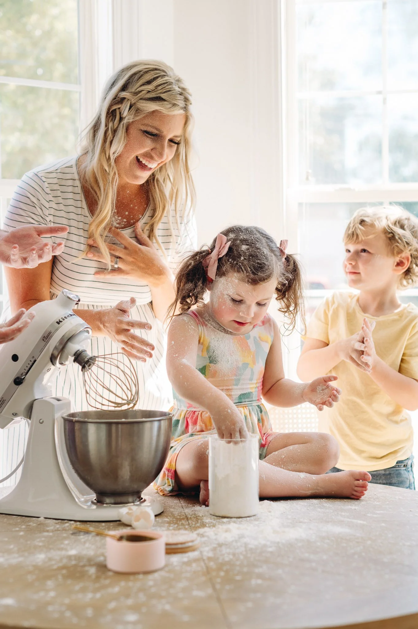 A woman and two children baking in a bright kitchen, with flour on their hands and counter, a mixer on the table, and large windows in the background, lifestyle family photograph in Bend, Oregon by Elise Souders Photography.