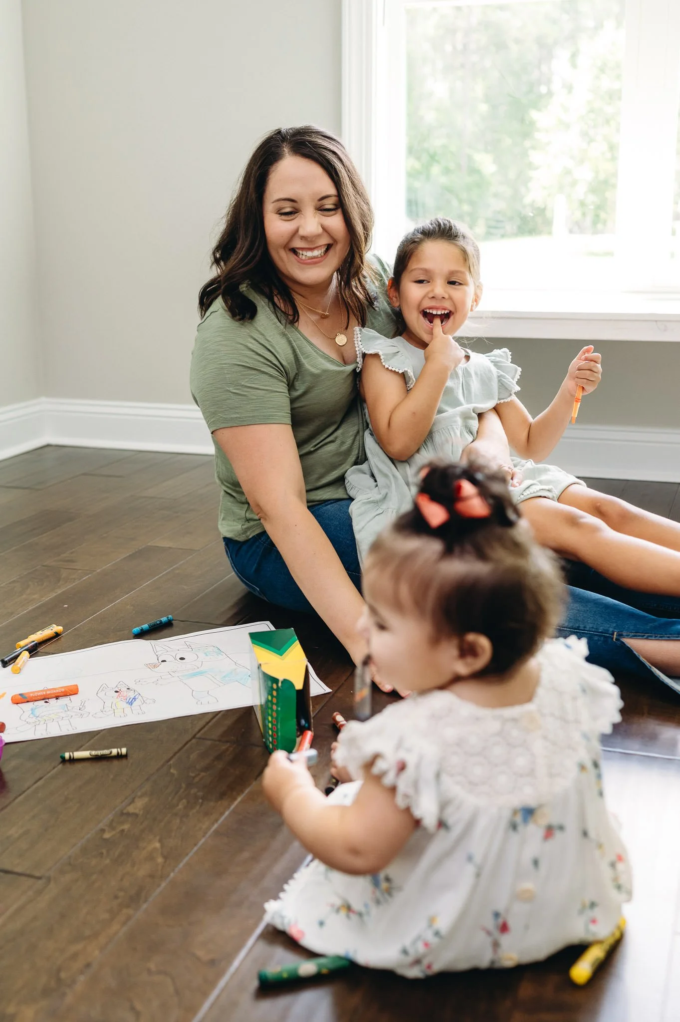 A woman and two young girls sitting on a wooden floor, playing with colored markers and drawings. The woman is smiling with closed eyes, wearing a green shirt, the girl to her right is laughing with a finger in her mouth and holding a marker, and the girl in the foreground has a bow in her hair and is focused on her drawing.