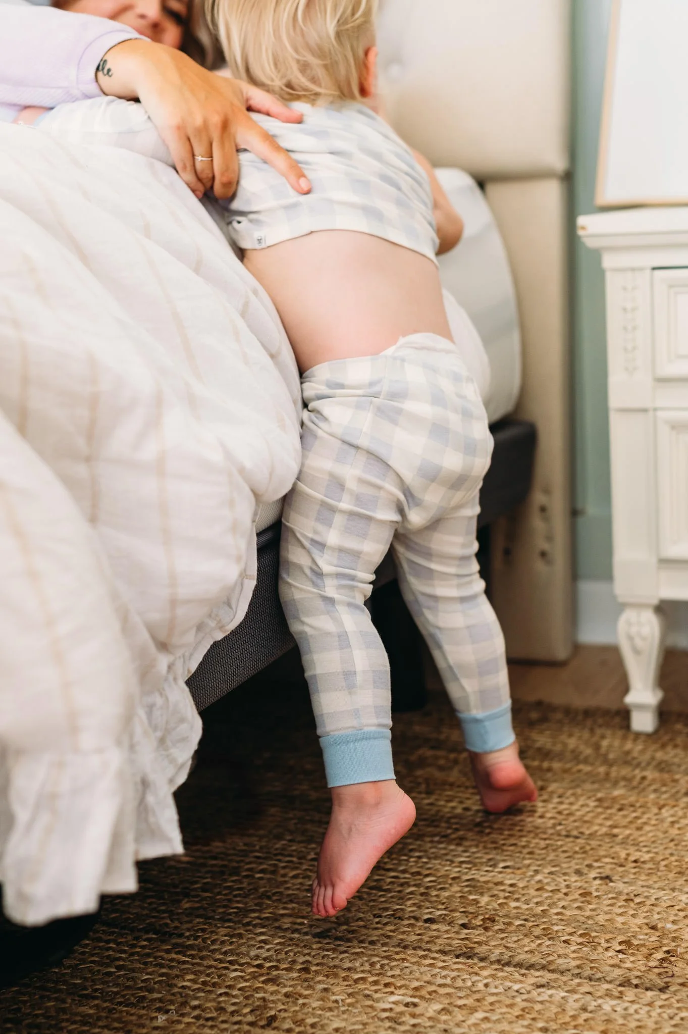 Child in pajamas standing on bedroom carpet, leaning over the side of a bed, with an adult hugging them from behind.