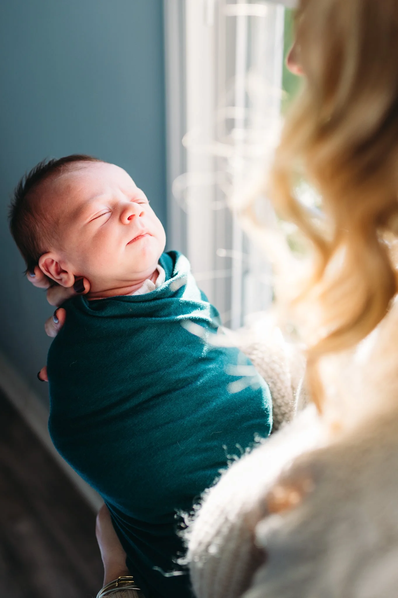 A woman holding a sleeping newborn baby near a window with sunlight coming through in a cozy lifestyle newborn session in-home in Bend, Oregon by Elise Souders.