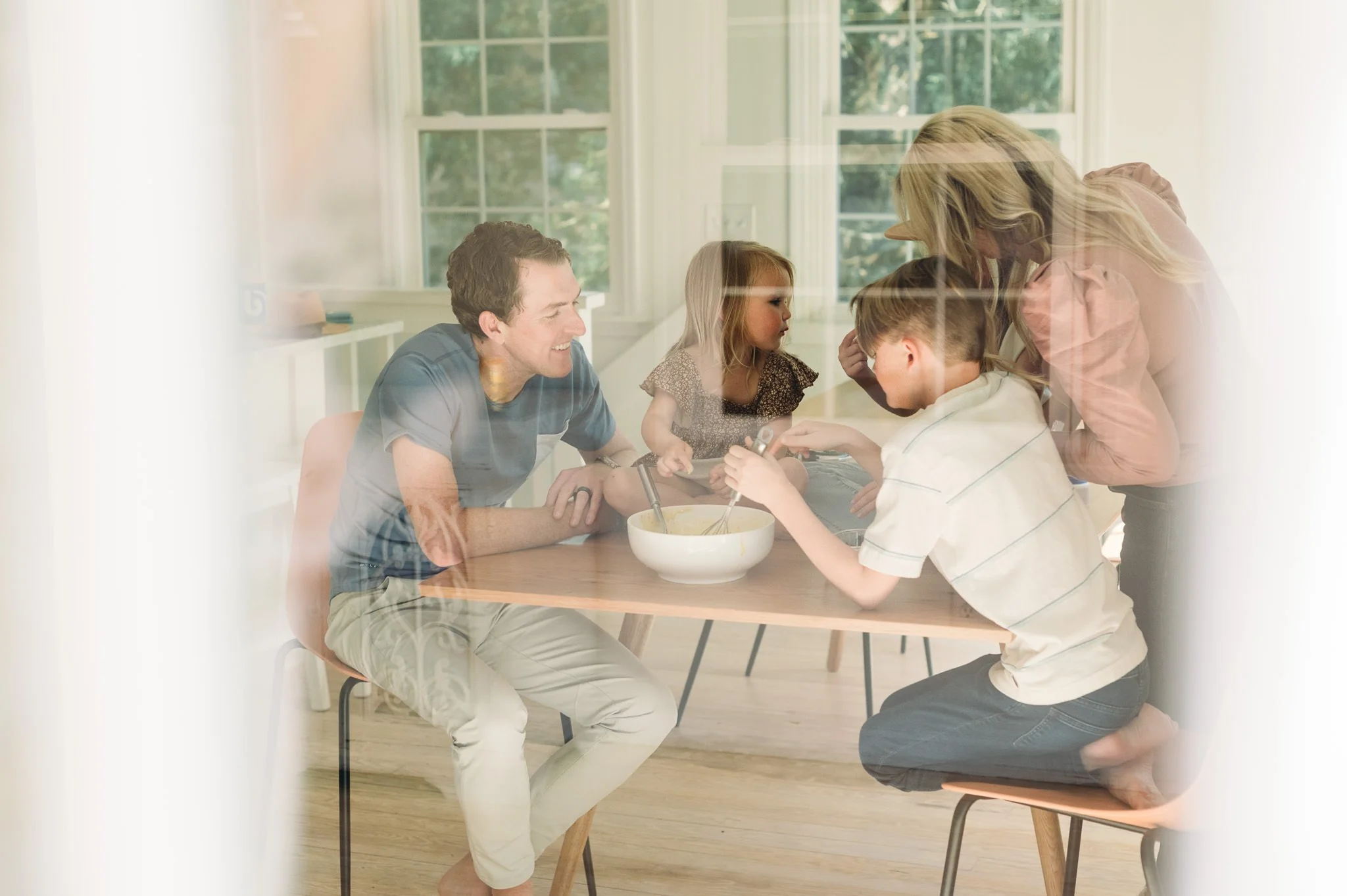 Family gathered around a table, baking together in a bright kitchen with large windows, lifestyle family photo taken from the perspective of outside the window by Elise Souders Photography