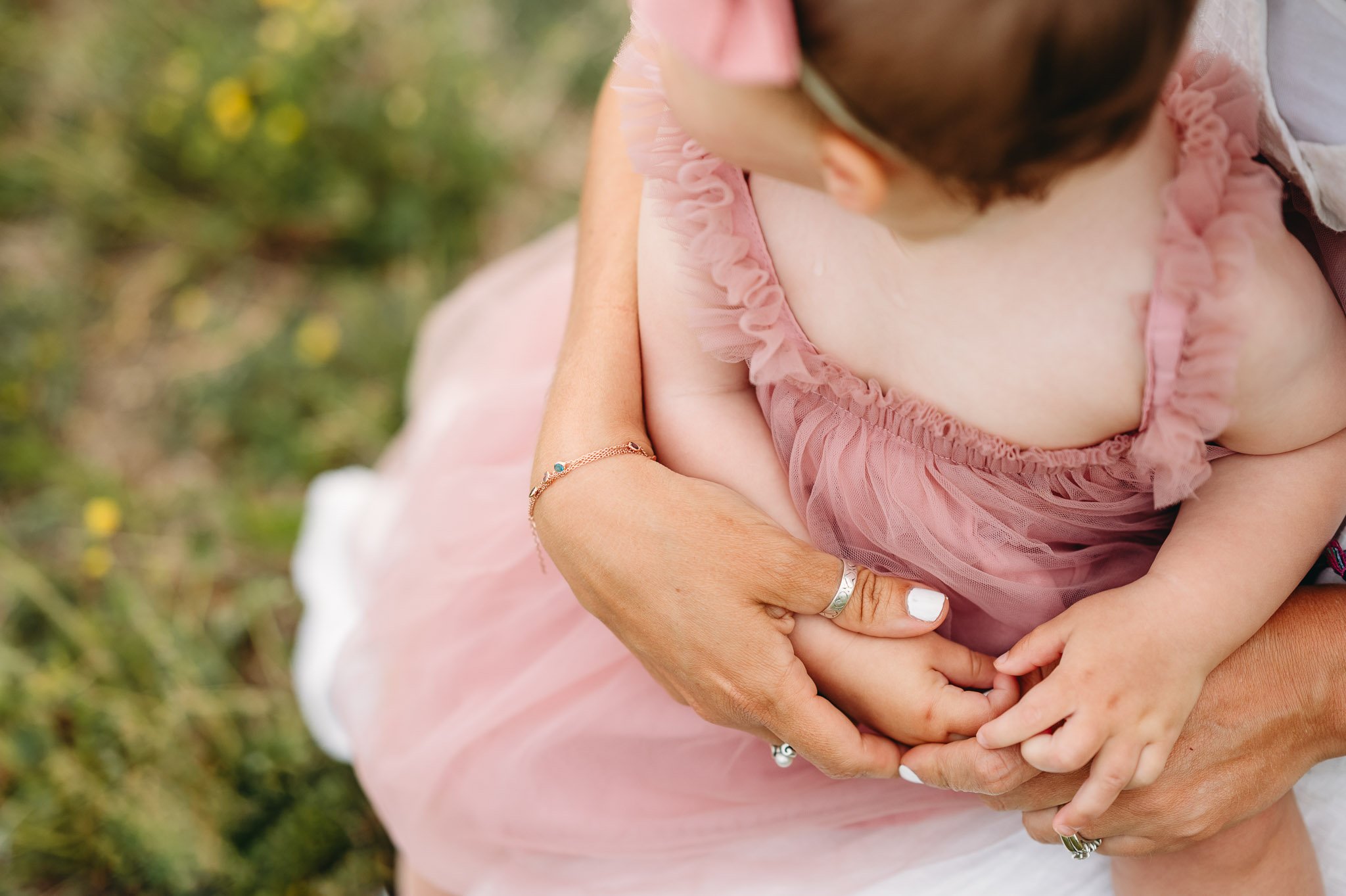 Detail image of mother's hands holding toddlers small fingers in a pink dress, sitting in a field on a summer's day.