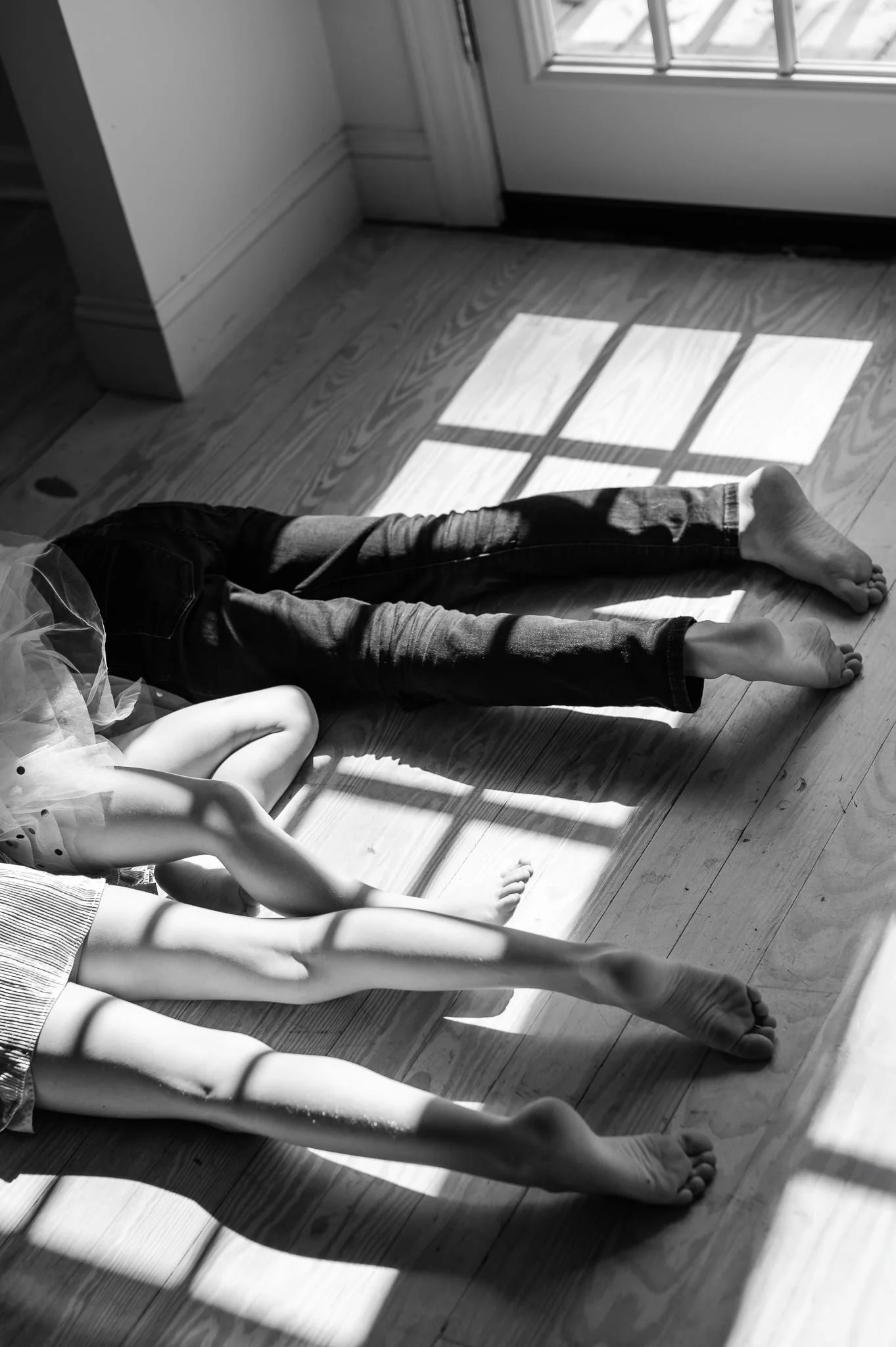 Black and white photo of three children lying on a floor with sunlight casting shadows through window panes. The children are barefoot, with part of a ballet tutu visible in the foreground in documentary family photo by Elise Souders Photography.