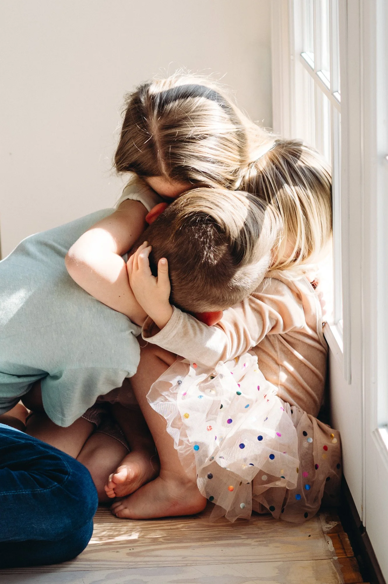 Mother hugging three young children beside a sunlit window