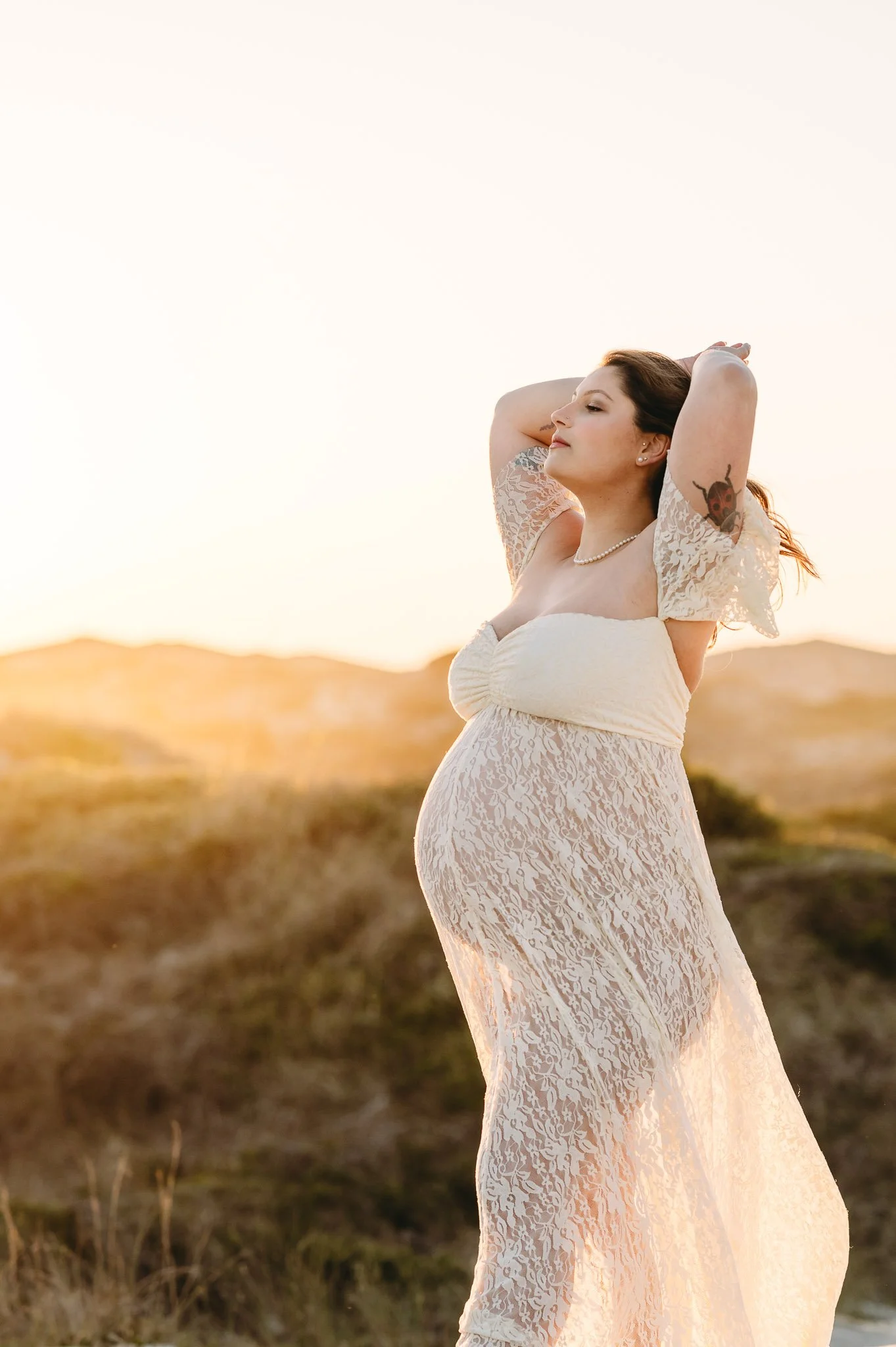 A woman in a white lace dress with puffed sleeves and a pearl necklace stands outdoors with her hands behind her head, eyes closed, in a warm, golden sunset landscape that illuminates her pregnant belly for a maternity photoshoot in Central Oregon