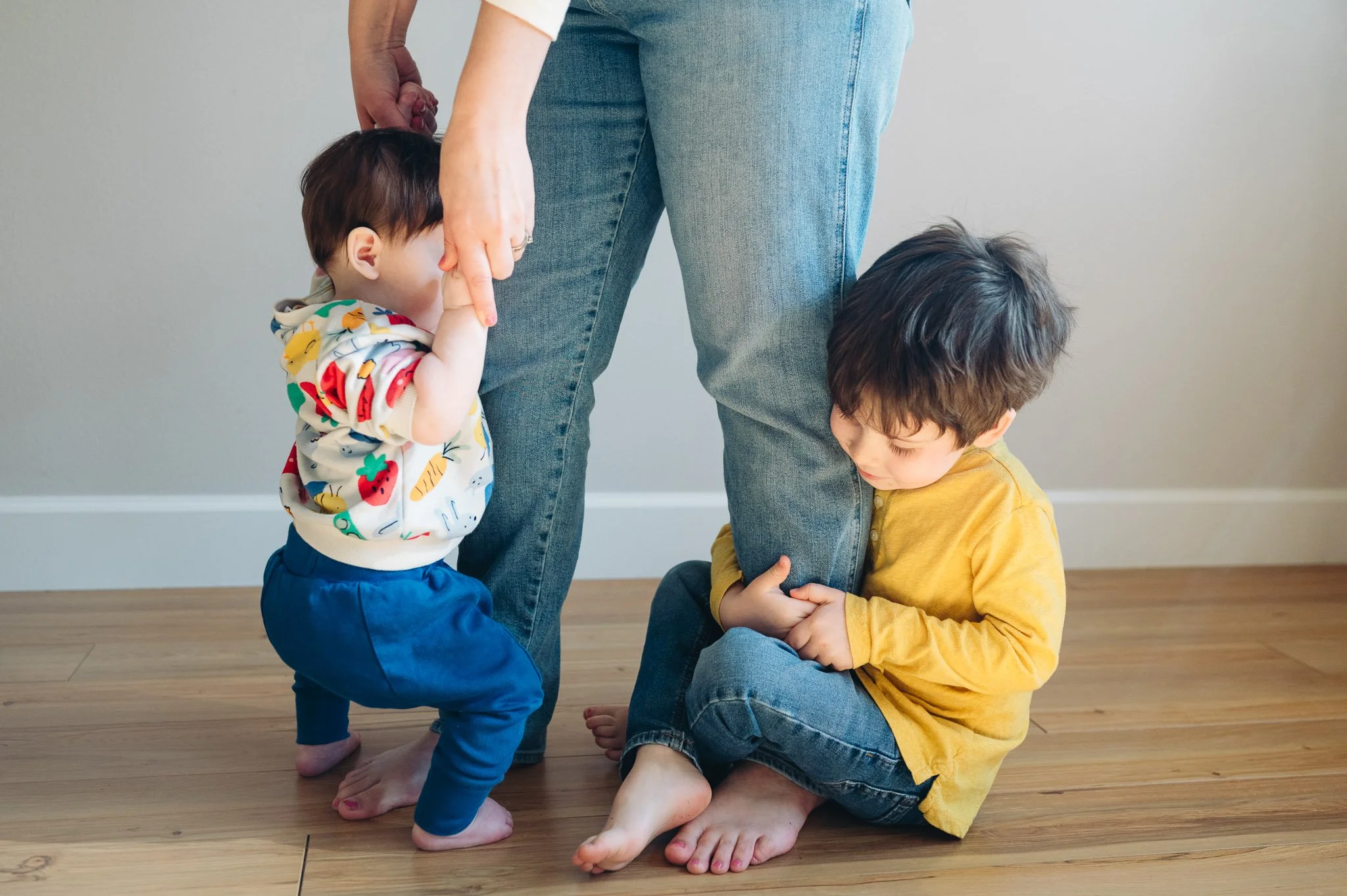 A woman helping a young girl and boy in a room with wooden floor, girl on her knees, holding the girl's hand, and boy sitting on the floor holding his ankle, both kids looking down.