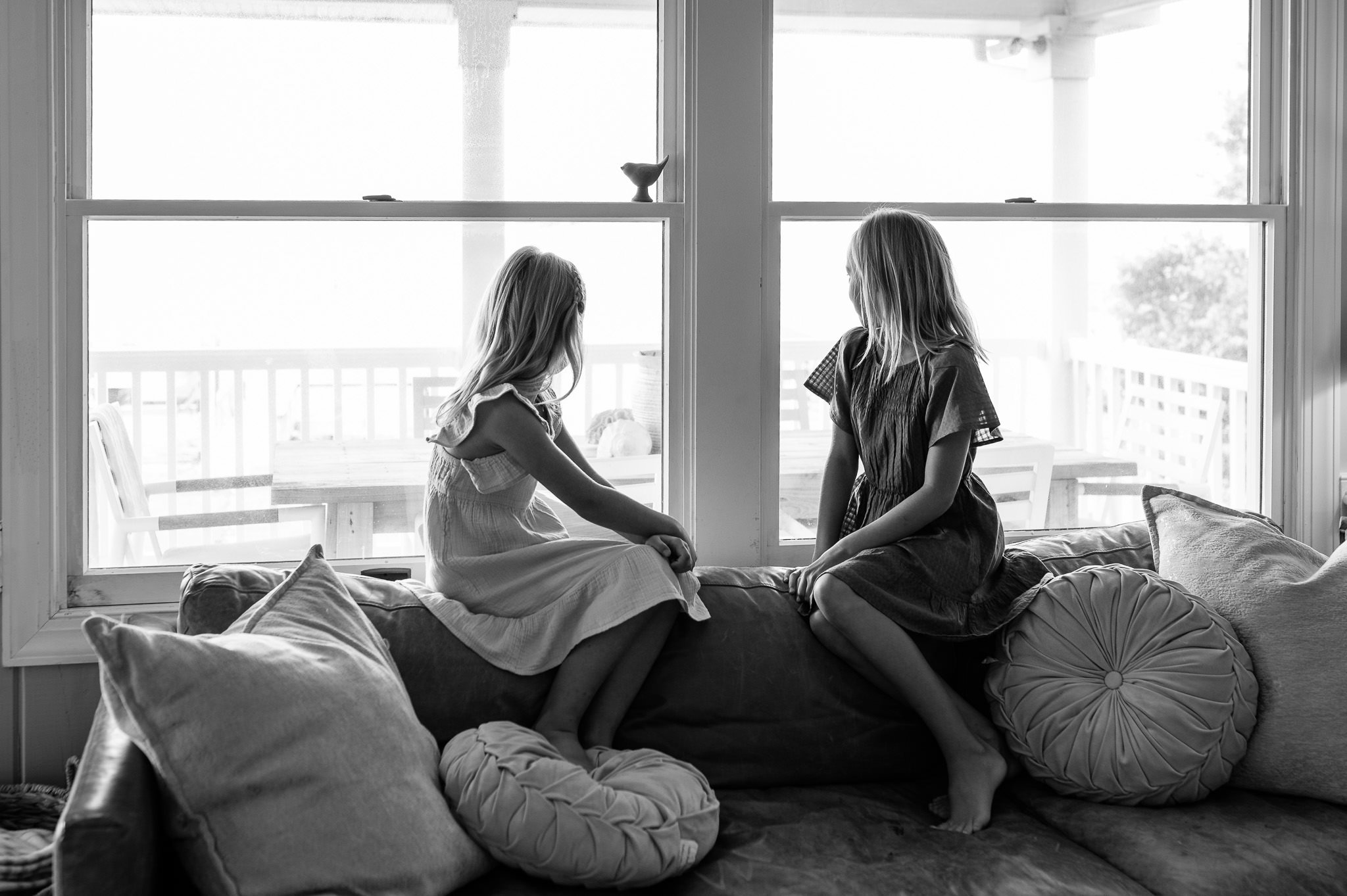Black and white image of two sisters looking outside their window while sitting on the couch, candid family photography in Bend, Oregon