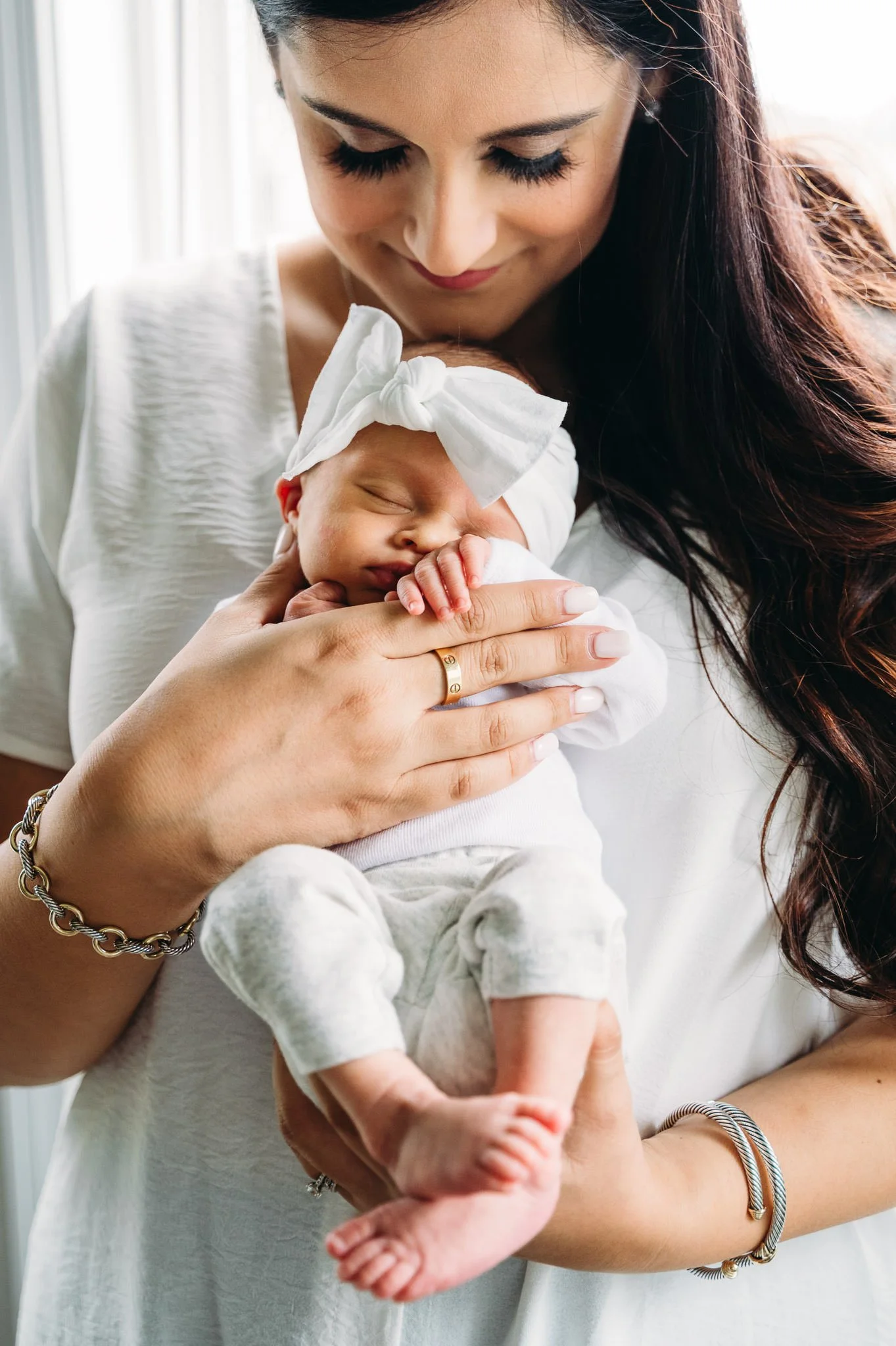 A woman with long dark hair and jewelry, wearing a white shirt, holding a sleeping baby in her arms. The baby is wearing a white outfit and a headband with a bow in a cozy newborn photoshoot in-home in Bend, Oregon