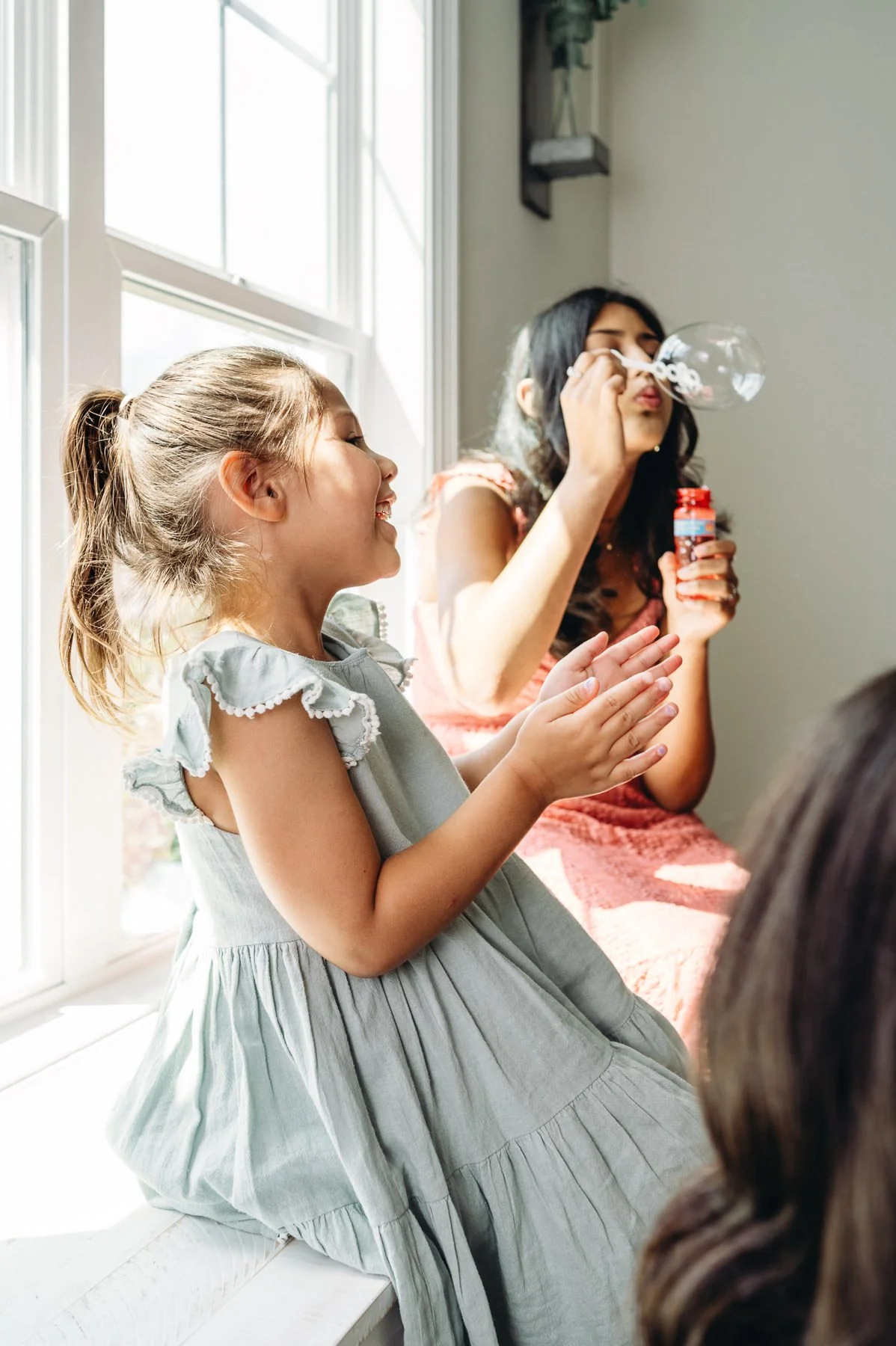 Sisters playing with bubbles in front of lit window, play-based session by Elise Souders Bend Family Photographer.